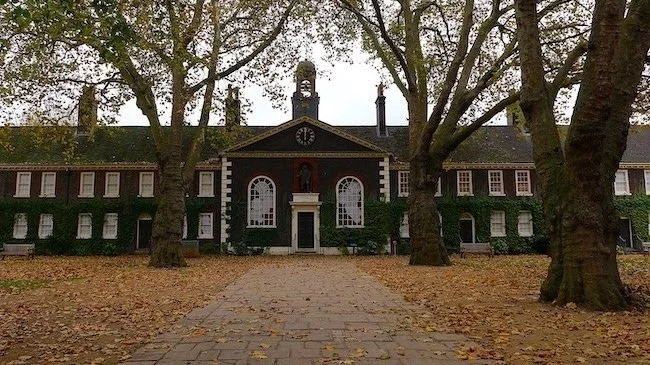 An old, dark-colored building with a clock on the front, surrounded by trees and fallen leaves, with a stone pathway leading to the entrance.