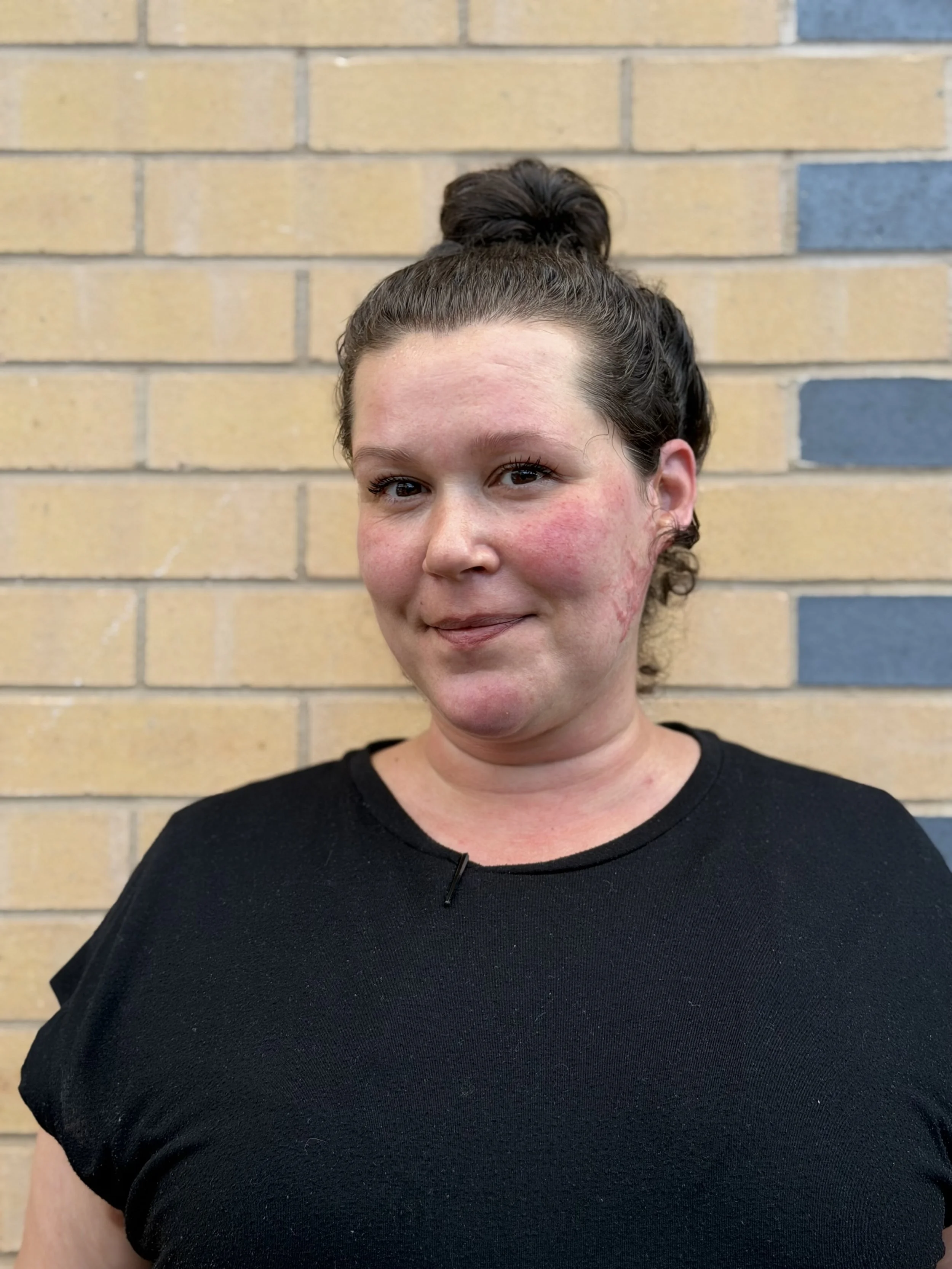A woman with dark hair in a bun, wearing a black shirt, is standing in front of a brick wall.