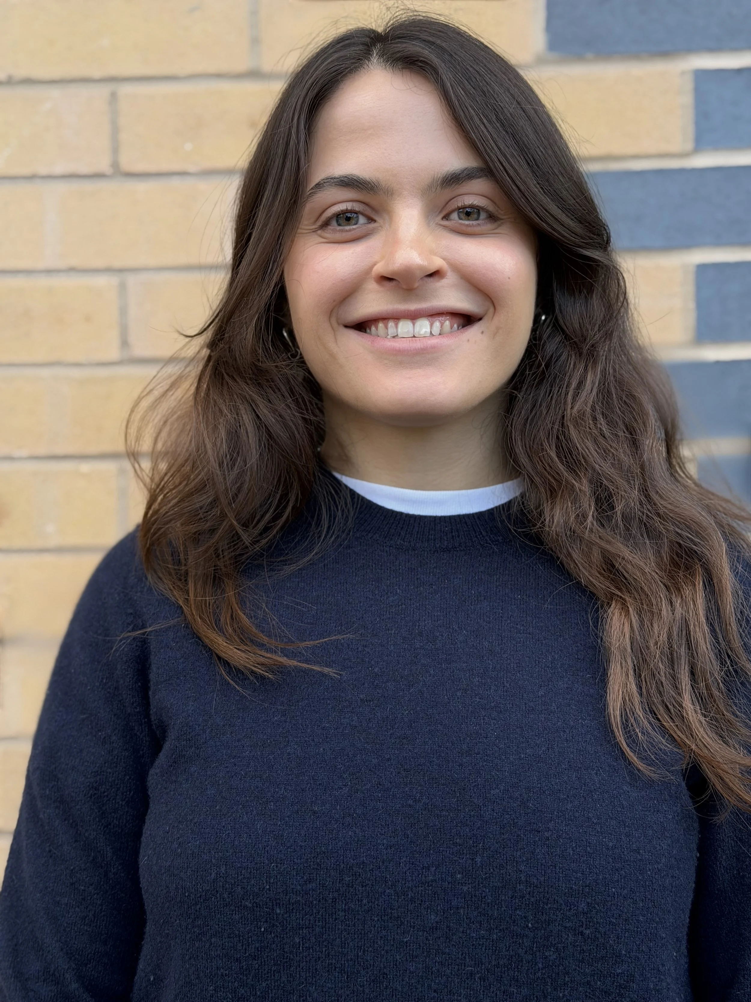 A young woman with long, wavy brown hair and light skin, smiling, wearing a navy blue sweater with a white collar, standing in front of a brick wall with yellow and blue bricks.