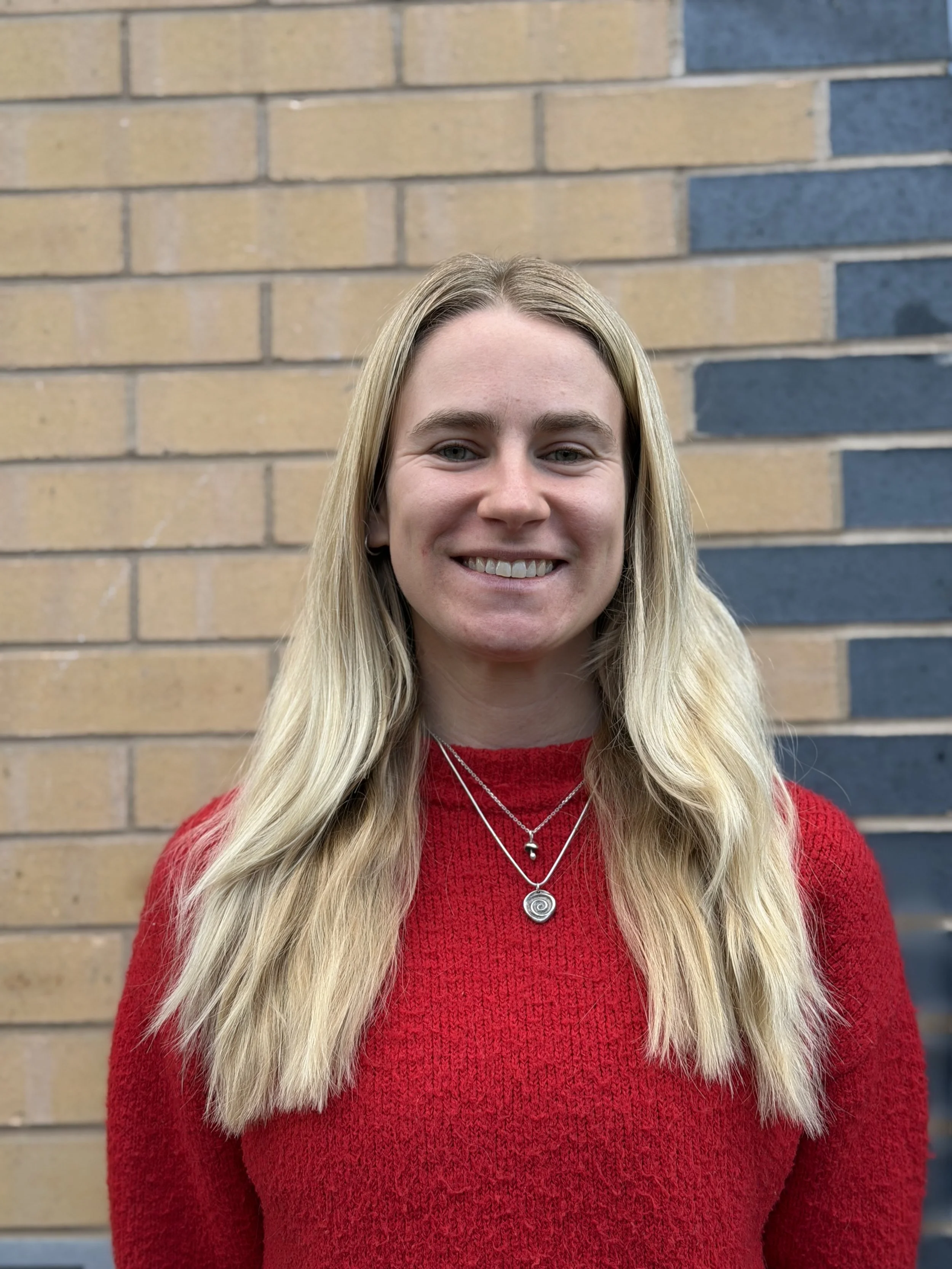 A woman with long blonde hair wearing a red sweater and layered necklaces, standing in front of a brick wall.