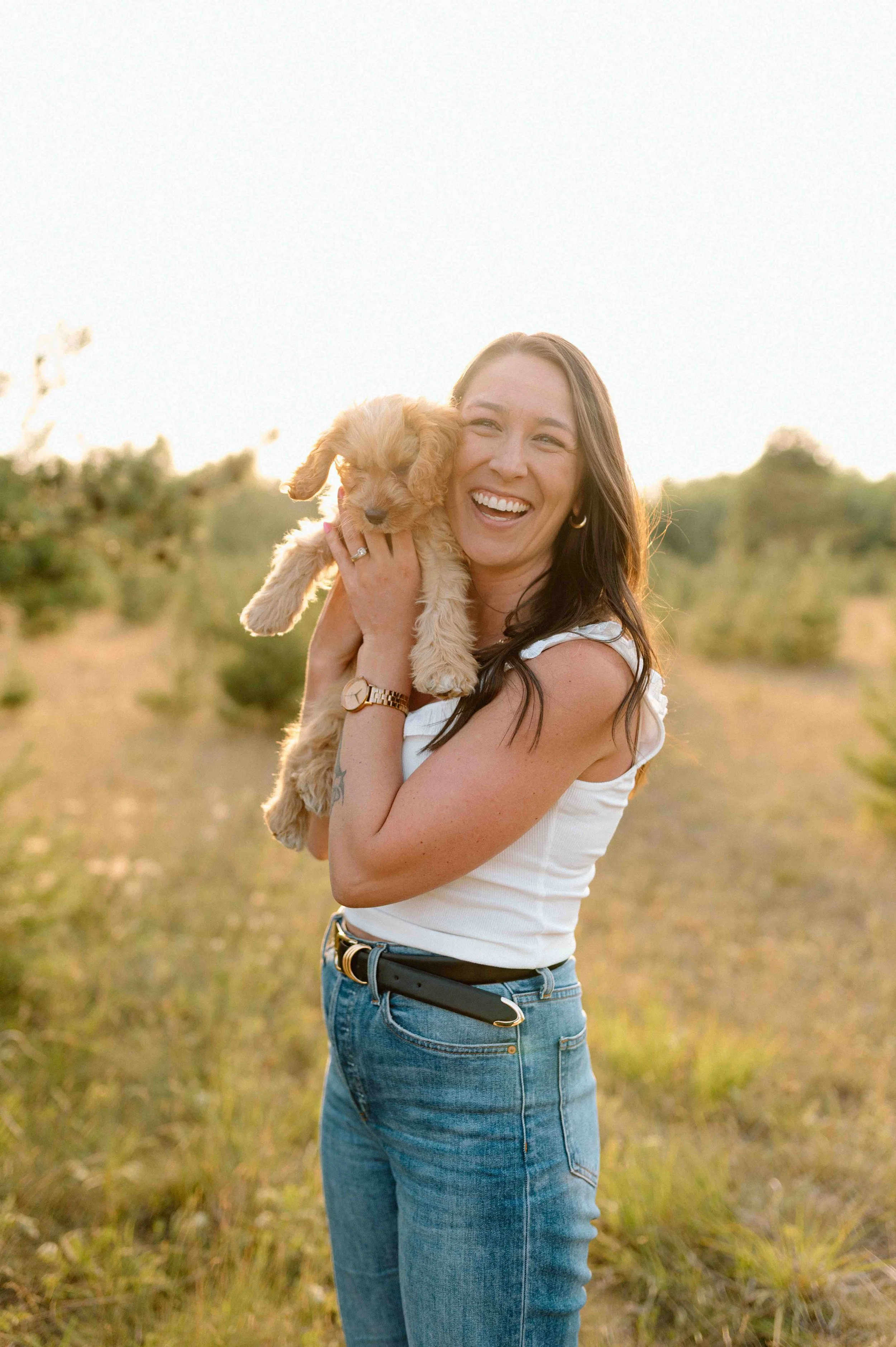 A woman with long dark hair, wearing a white sleeveless top and blue jeans, is smiling and holding a small, fluffy tan puppy in an outdoor field during sunset.