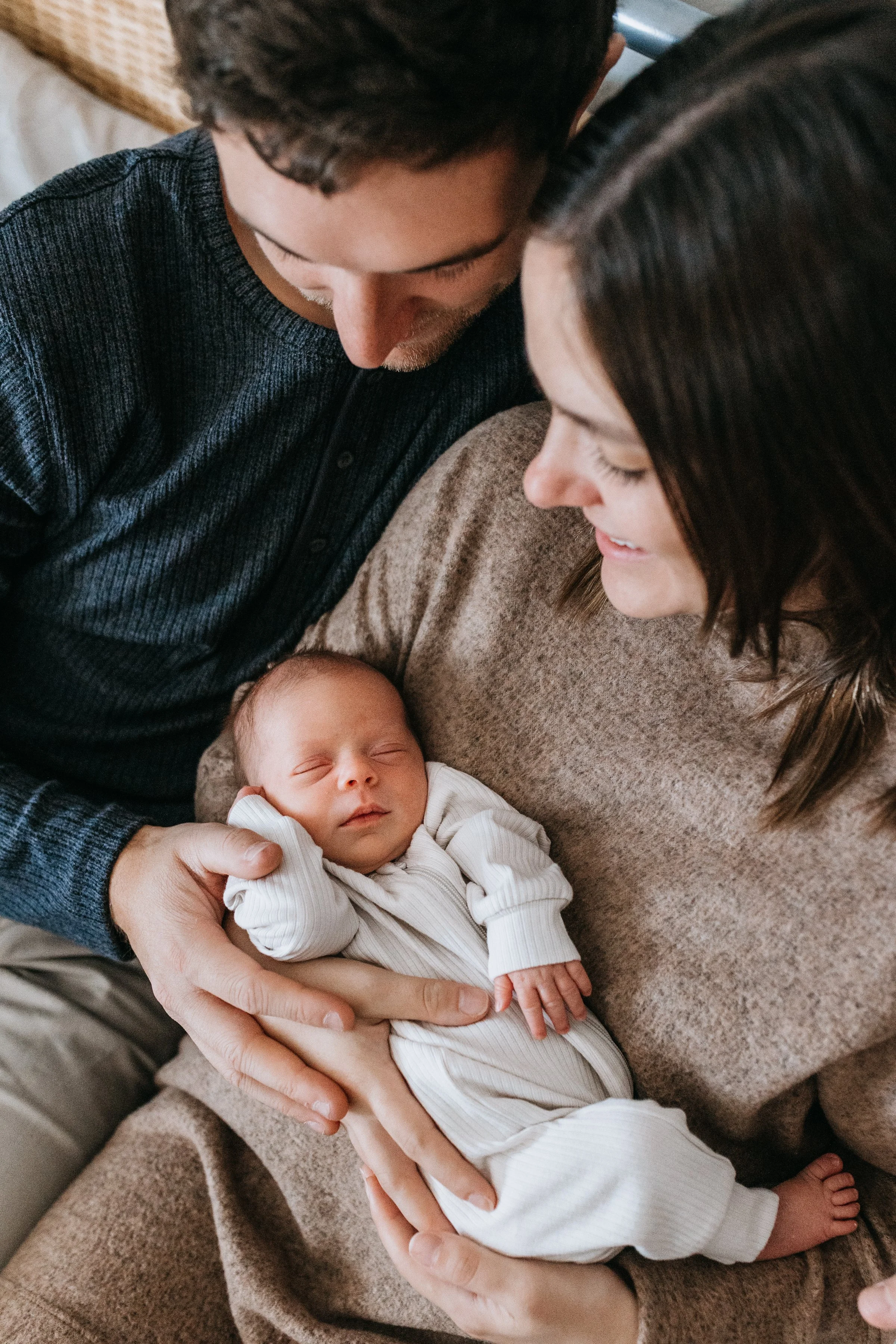 A man and a woman are holding a sleeping baby together, looking at the baby lovingly. They are sitting on a sofa with a textured blanket, and the scene feels warm and intimate.