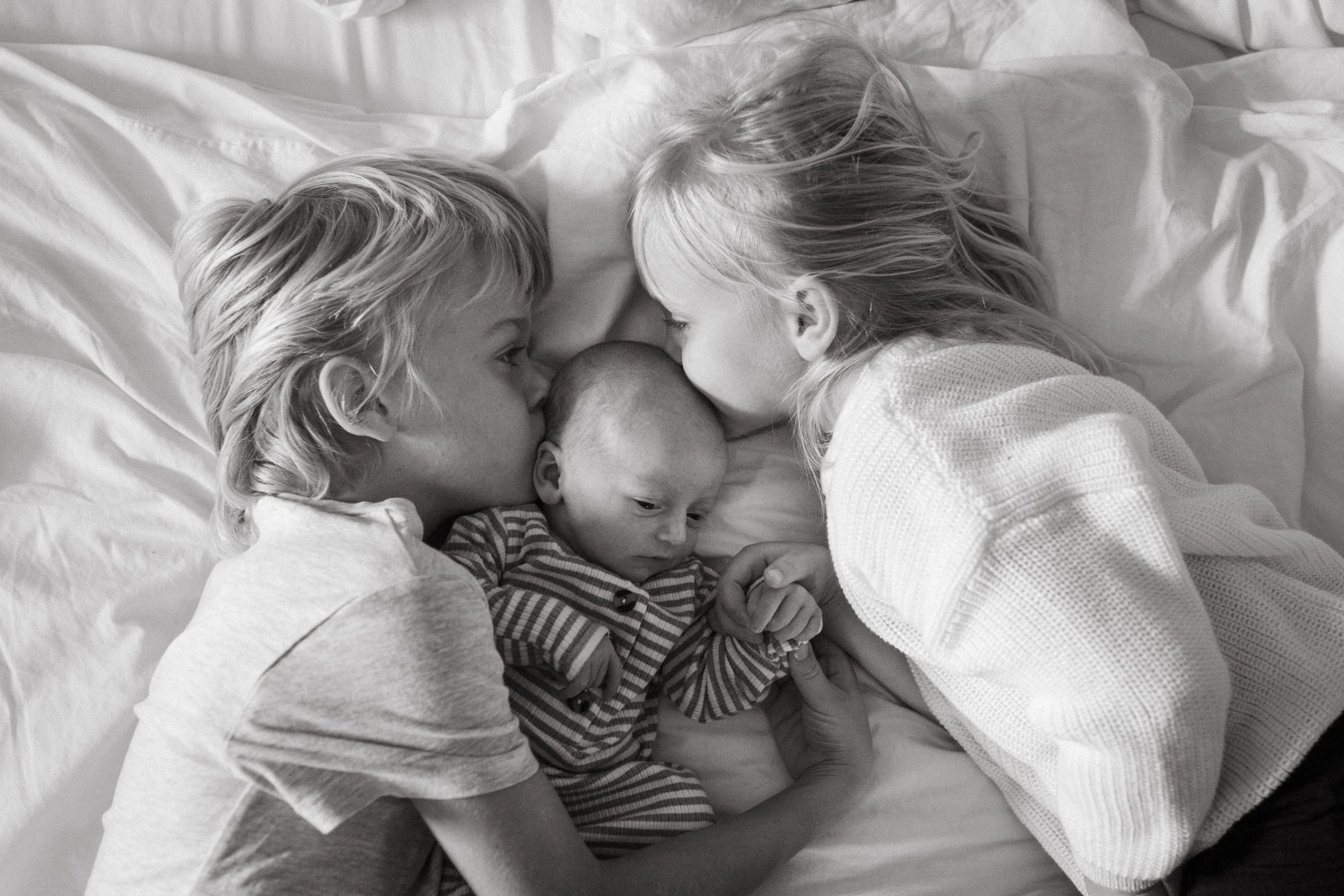 A family of three young children, two girls and a boy, lying closely together on a bed, touching foreheads, sharing an intimate moment in black and white.