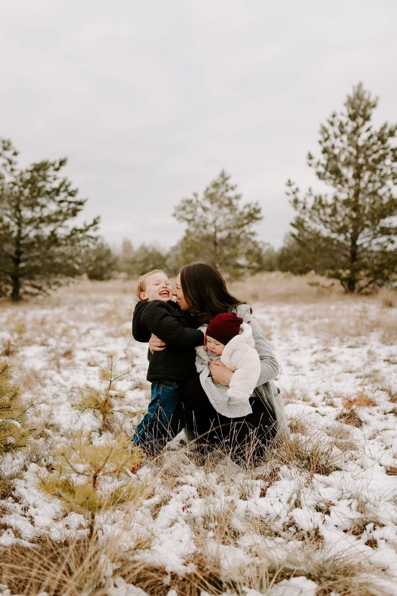A woman and two children, one girl and one baby, smiling and hugging outdoors in a snowy field with pine trees and overcast sky in the background.