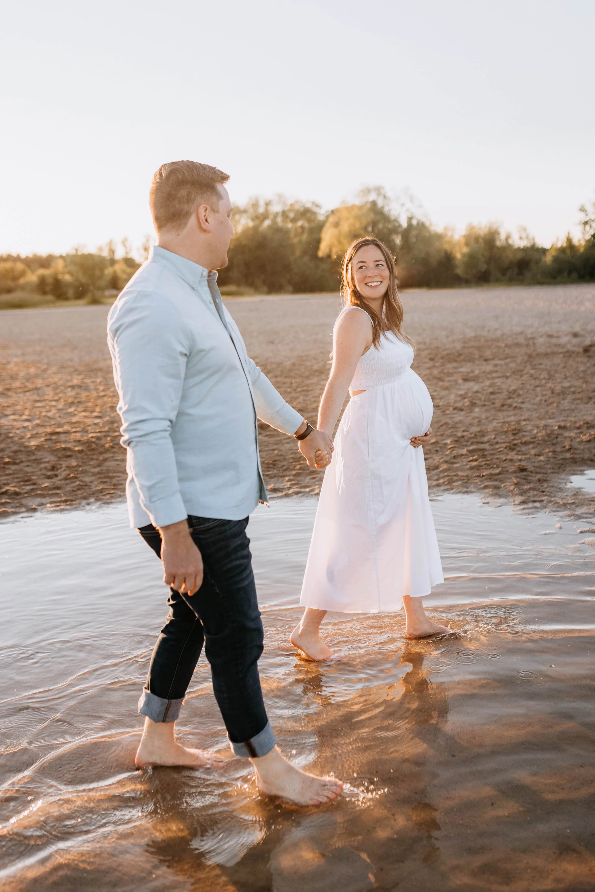A pregnant woman and a man are walking barefoot at the beach during sunset, holding hands with the woman smiling at the man.