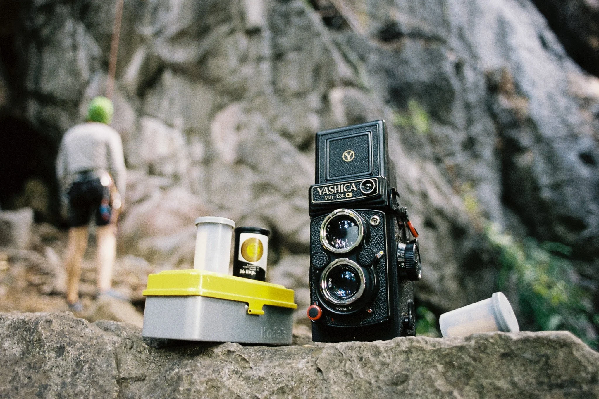 A vintage Yashica camera placed on a rocky surface along with film bottles and a yellow container. In the background, a person with a green hat is walking away, on a trail passed a rocky, natural environment.