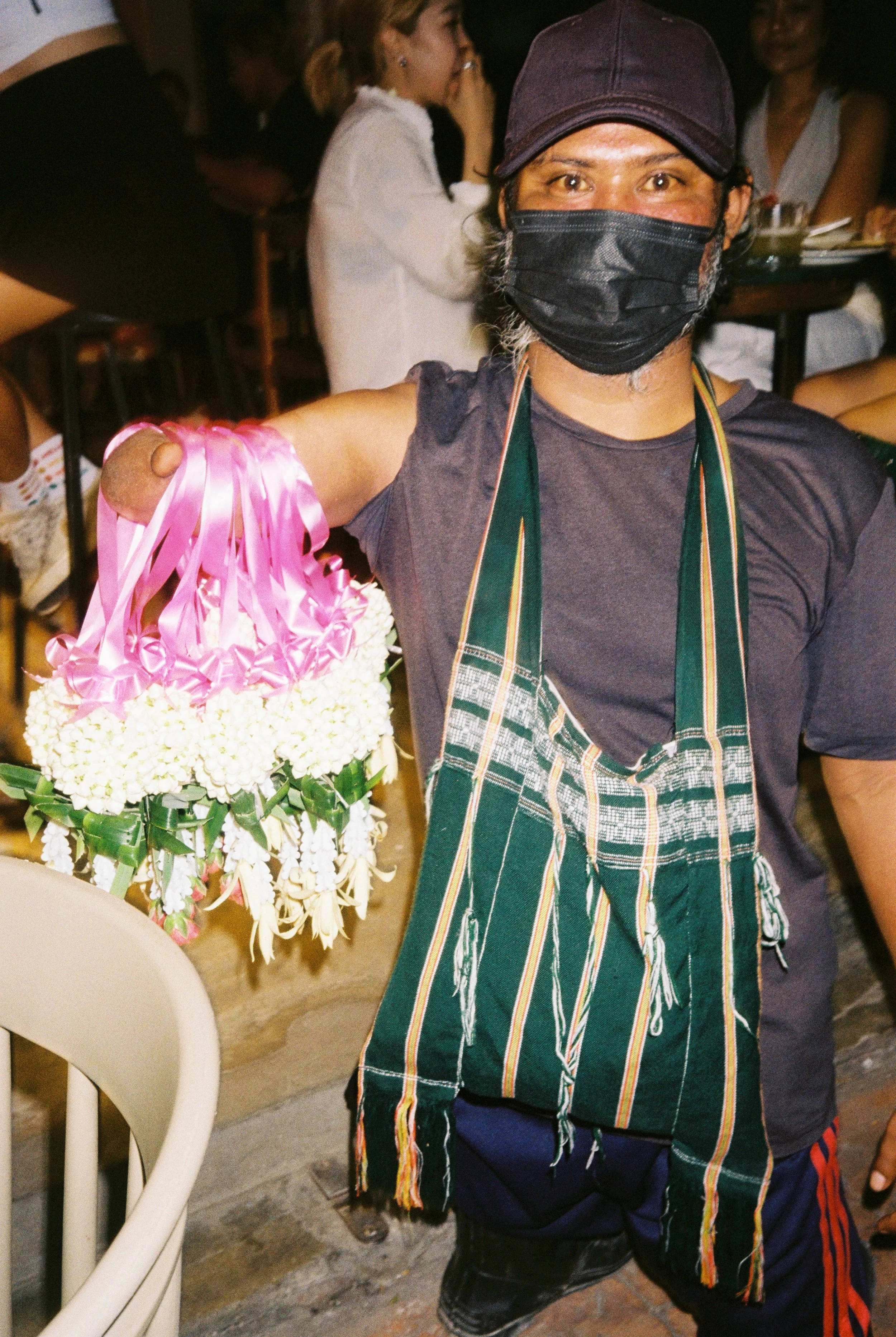 A Thai flower salesman wearing a face mask and a purple cap is holding a floral arrangement with white flowers and pink ribbons, standing indoors with several people in the background.