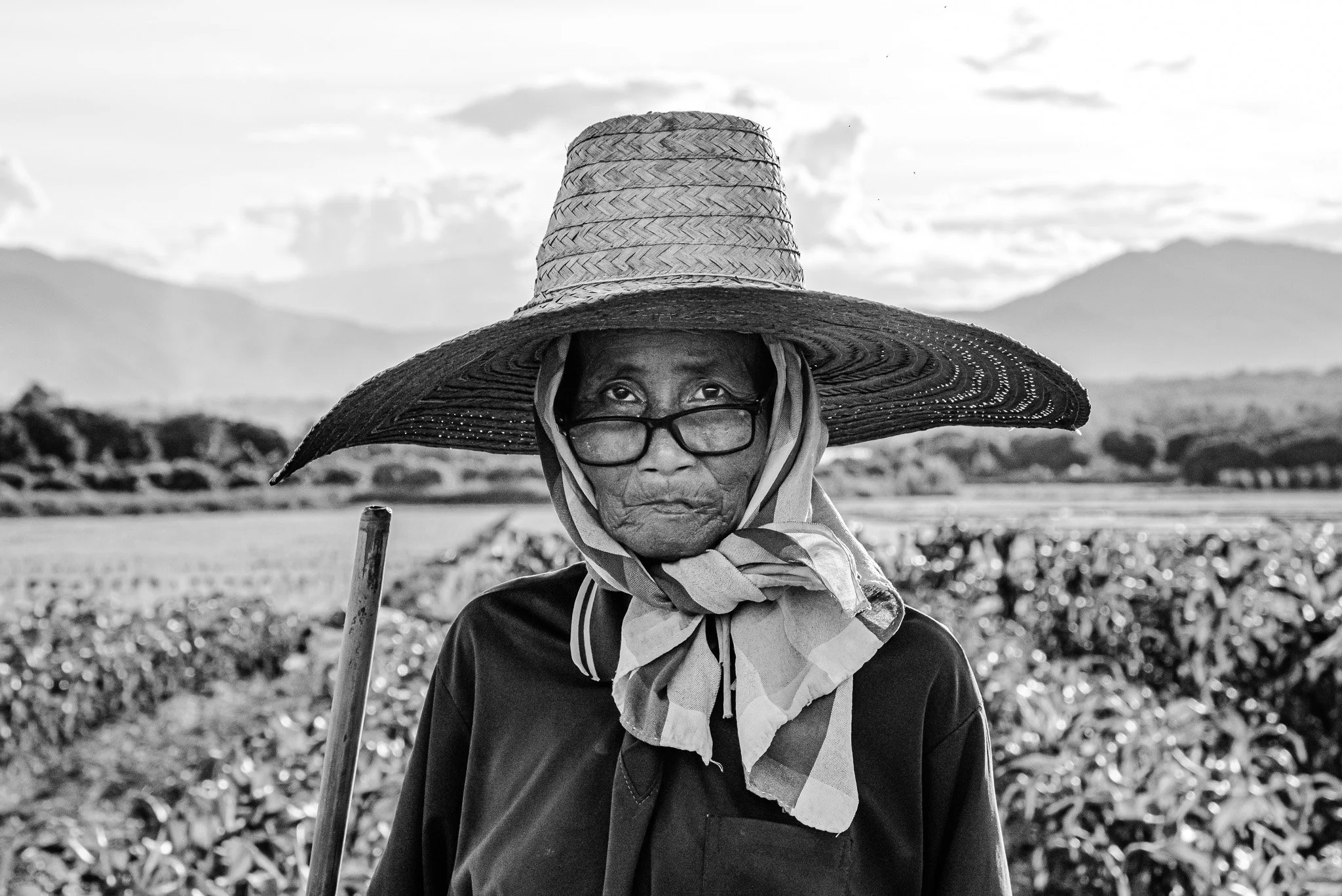 An elderly woman wearing a large woven sun hat, glasses, a scarf, and dark clothing, standing in a field with mountainous landscape in the background, black and white photograph.