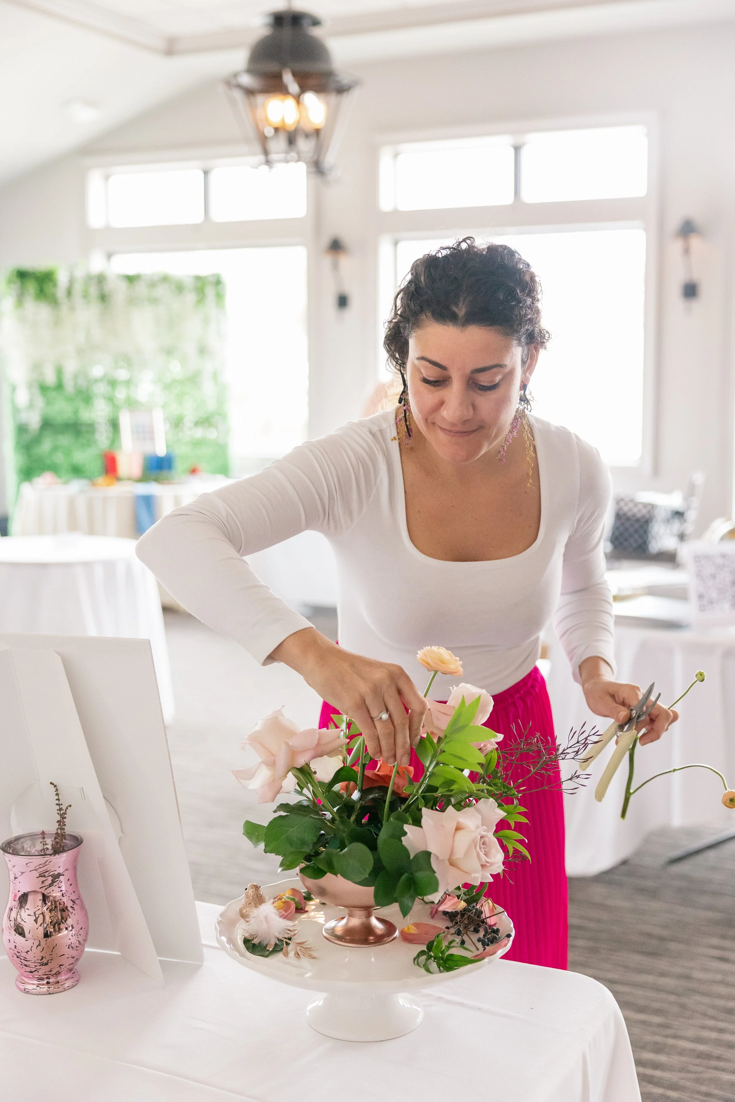 Woman arranging pink and peach flowers in a vase at a table, with scissors in her right hand, in a bright room with large windows.
