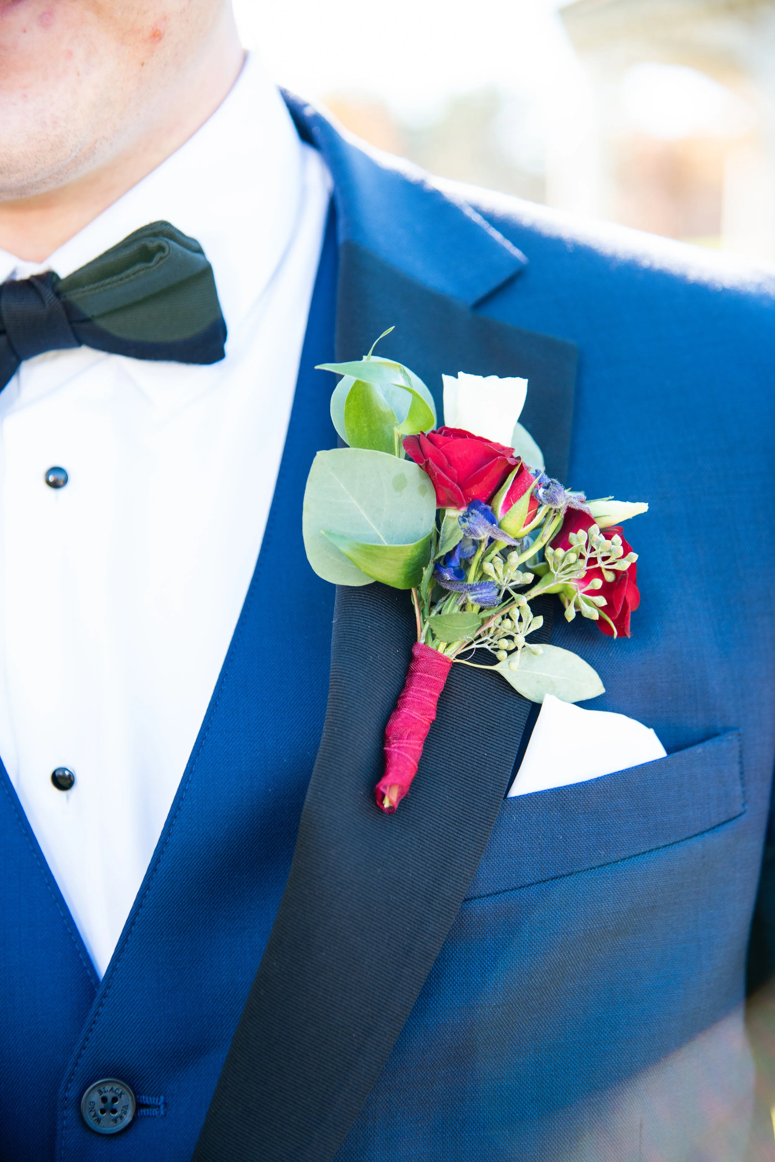Close-up of a man in a blue tuxedo with a black bow tie, wearing a red boutonniere with roses and greenery on his lapel.