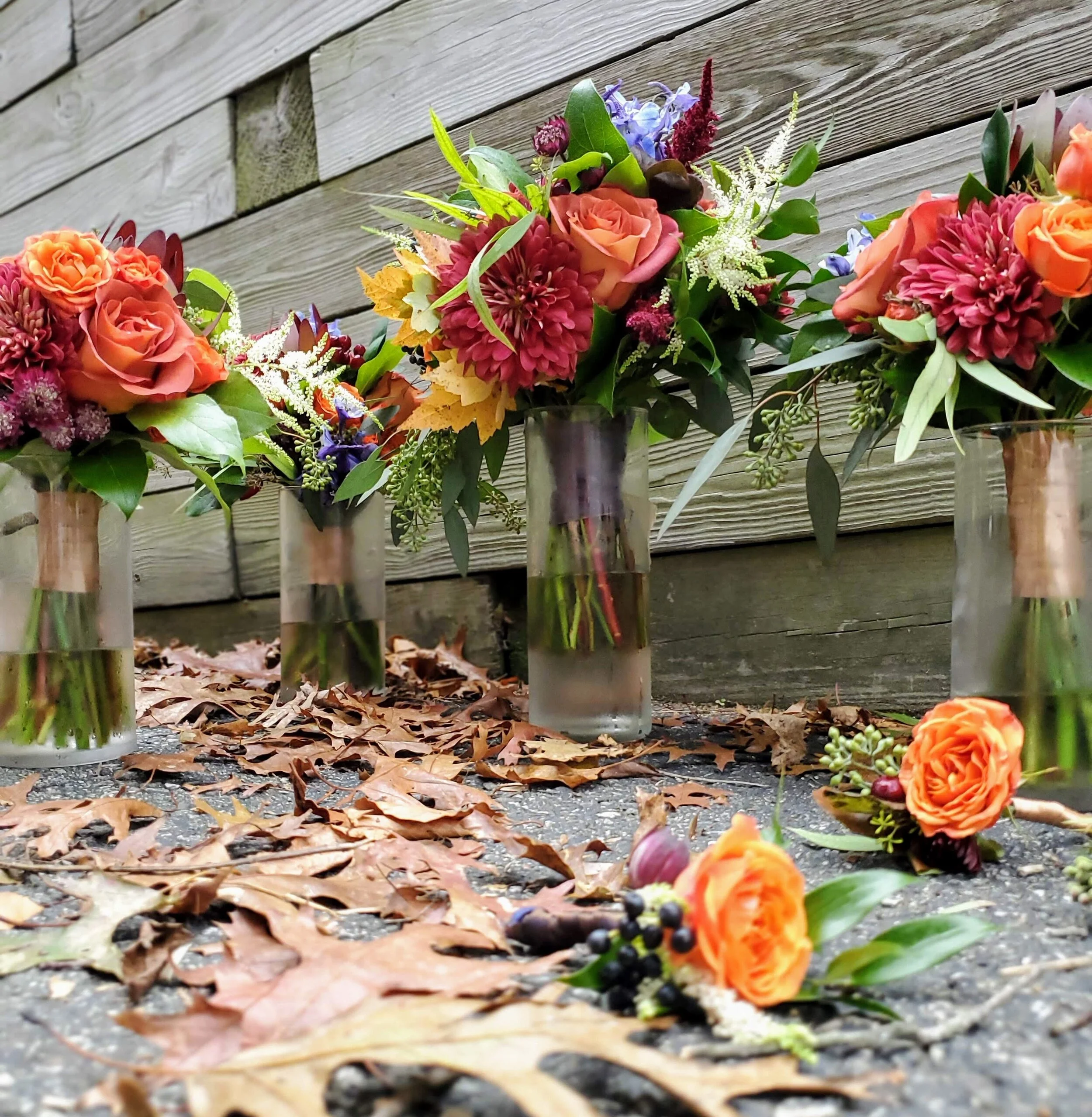 Four vases with colorful autumn flower arrangements on a surface covered with fallen leaves and berries, against a wooden background.