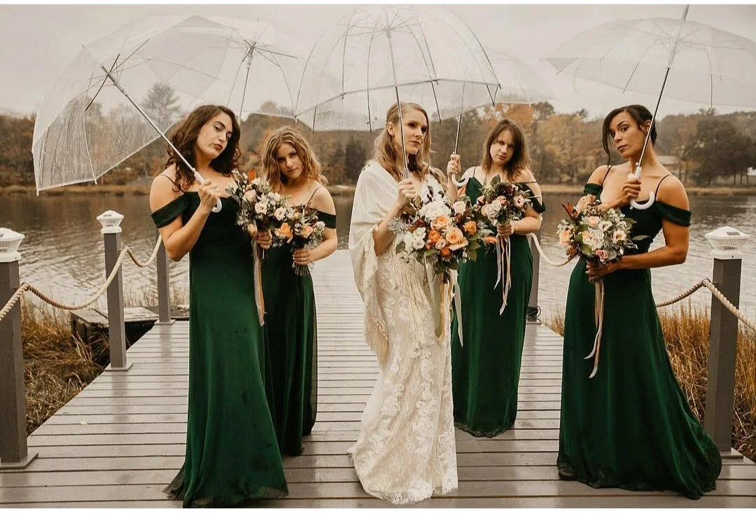A bride and four bridesmaids standing on a wooden dock by a lake, holding bouquets and clear umbrellas, dressed in formal green dresses, with trees in the background on a cloudy day.