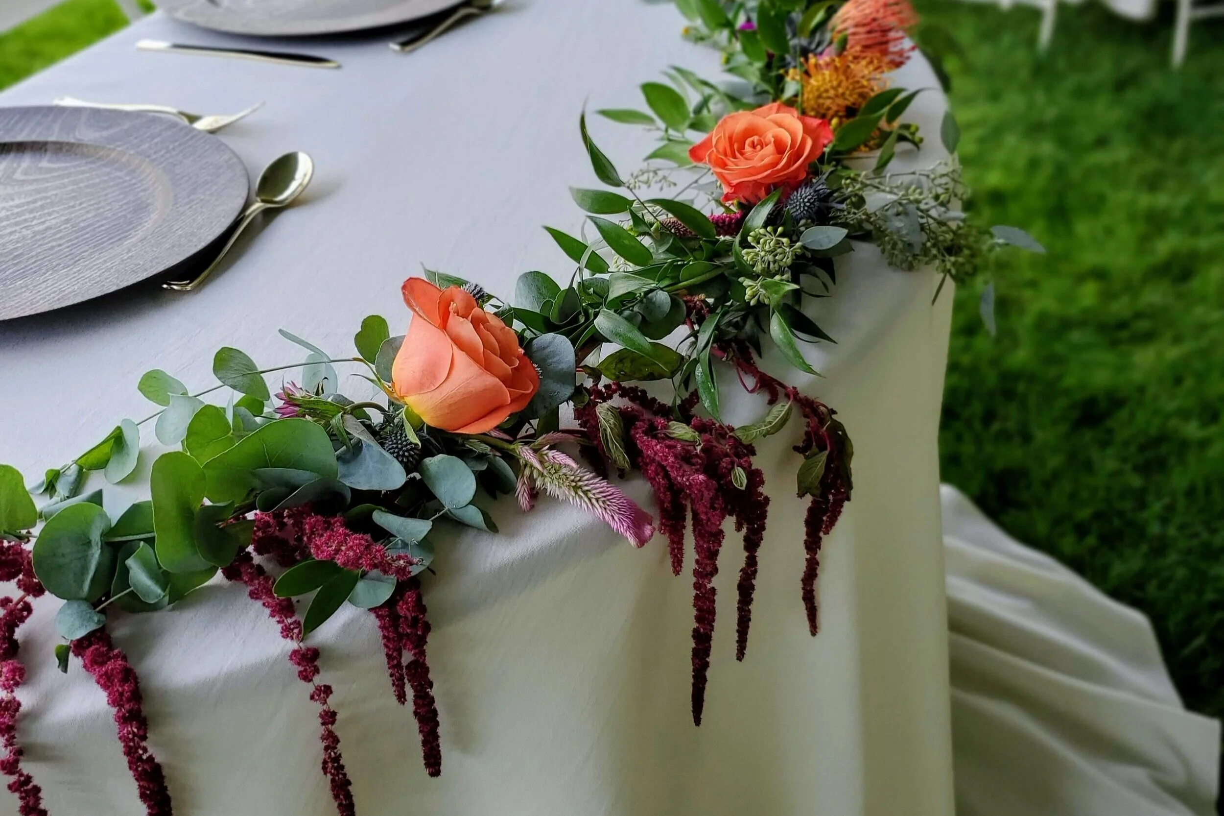 Decorative table runner with orange roses, green foliage, and hanging maroon flowers on a white tablecloth, set for an outdoor event.
