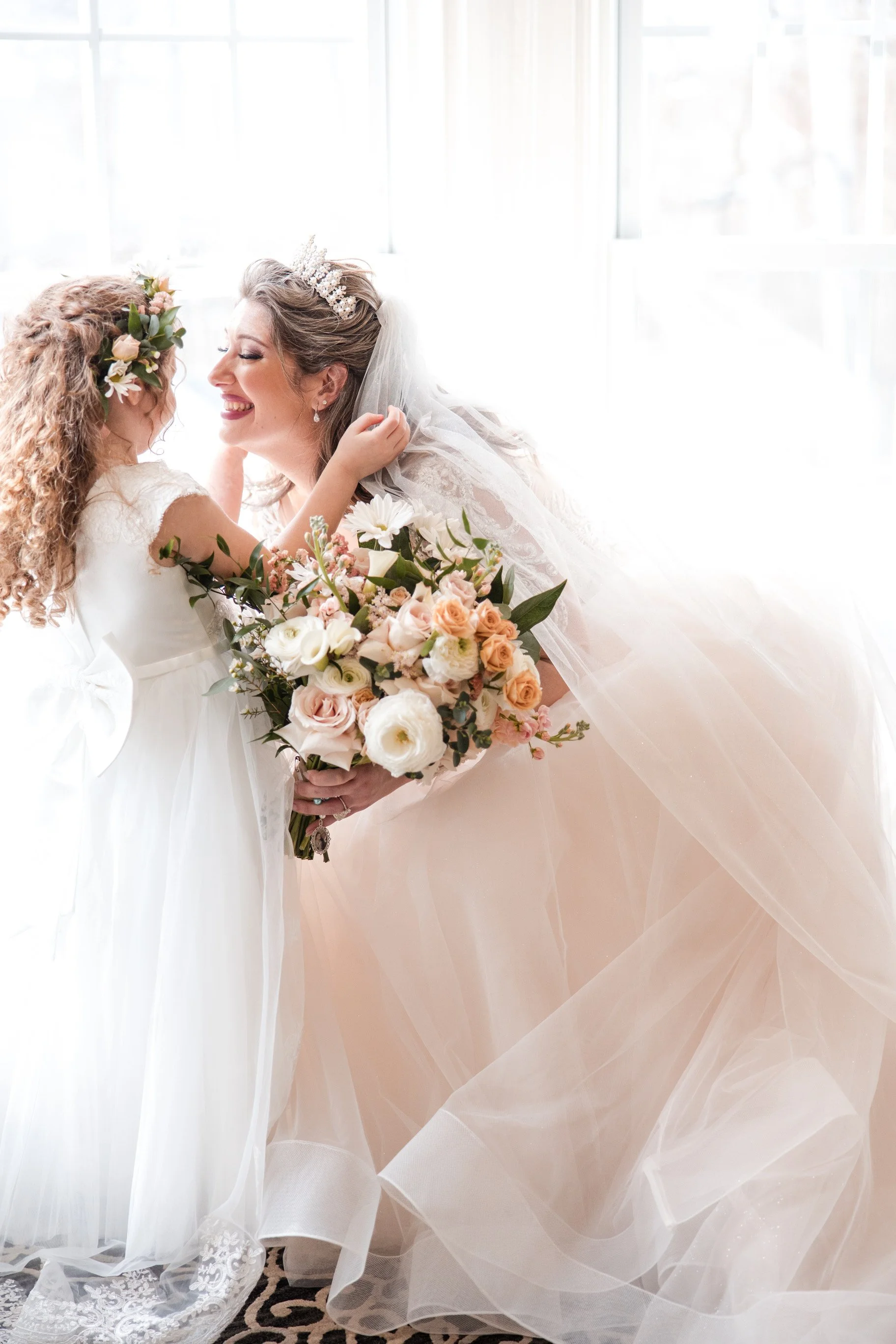 A bride holding a bouquet of flowers, smiling, interacting with a young girl with curly hair and a flower crown, in a bright room with sheer curtains.