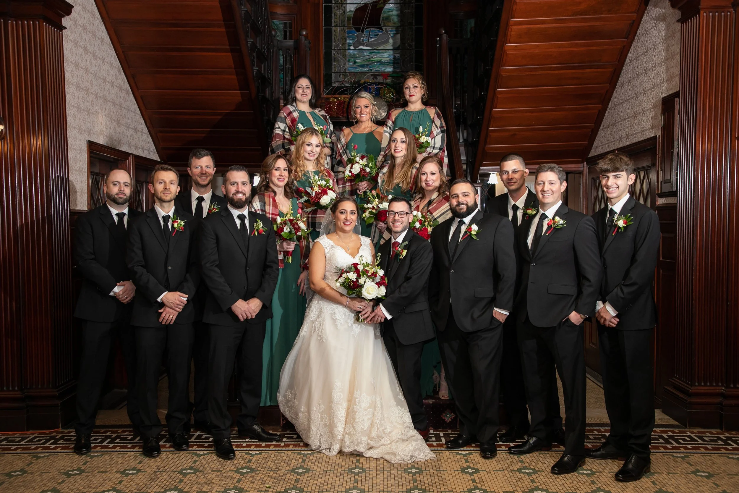 Group photo of a wedding party with bride and groom in the center, surrounded by bridesmaids holding bouquets and groomsmen, inside a wood-paneled venue with stained glass window and staircase.