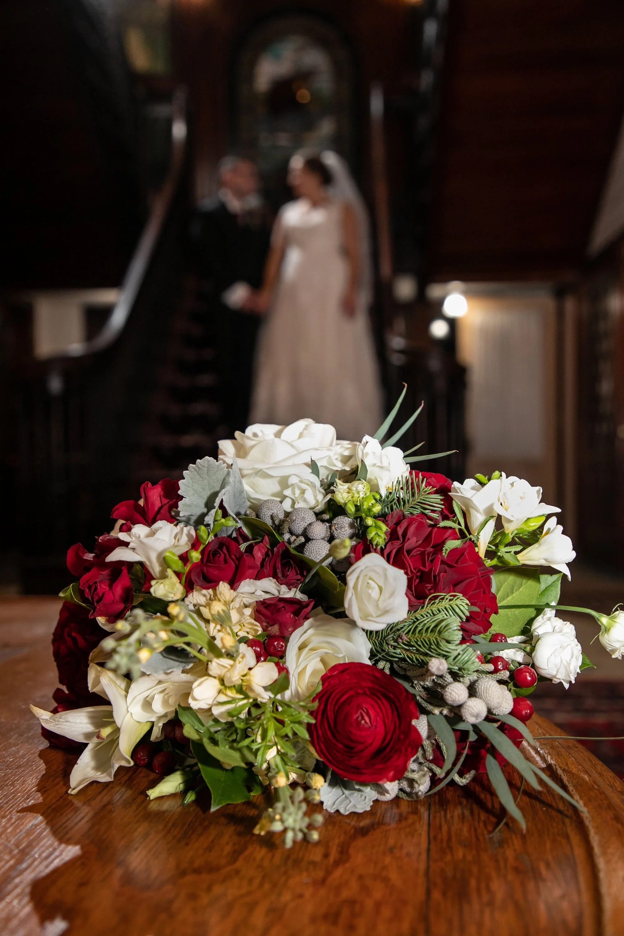 A wedding bouquet with red, white, and green flowers on a wooden table, with a bride and groom blurred in the background.