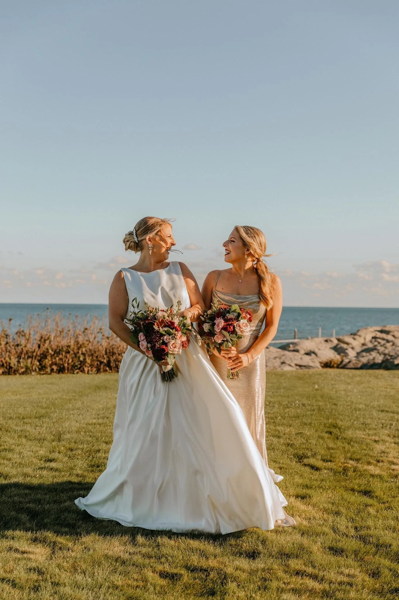 Two women, one in a wedding dress and the other in a gold gown, holding bouquets and smiling at each other outdoors near the water under a clear sky.