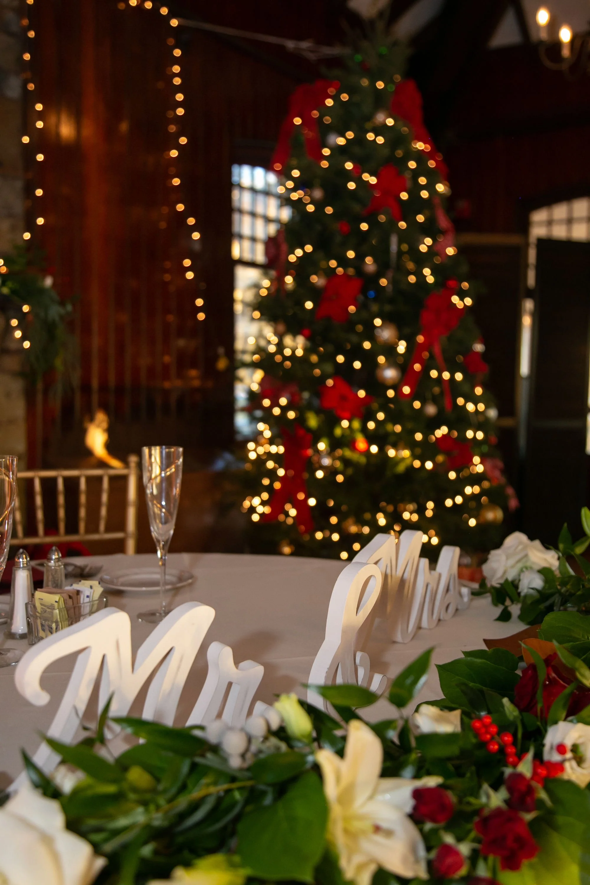 Decorated Christmas tree with red bows and ornaments inside a cozy wooden room, with a dining table decorated with a floral arrangement and white 'Mr. & Mrs.' signs.