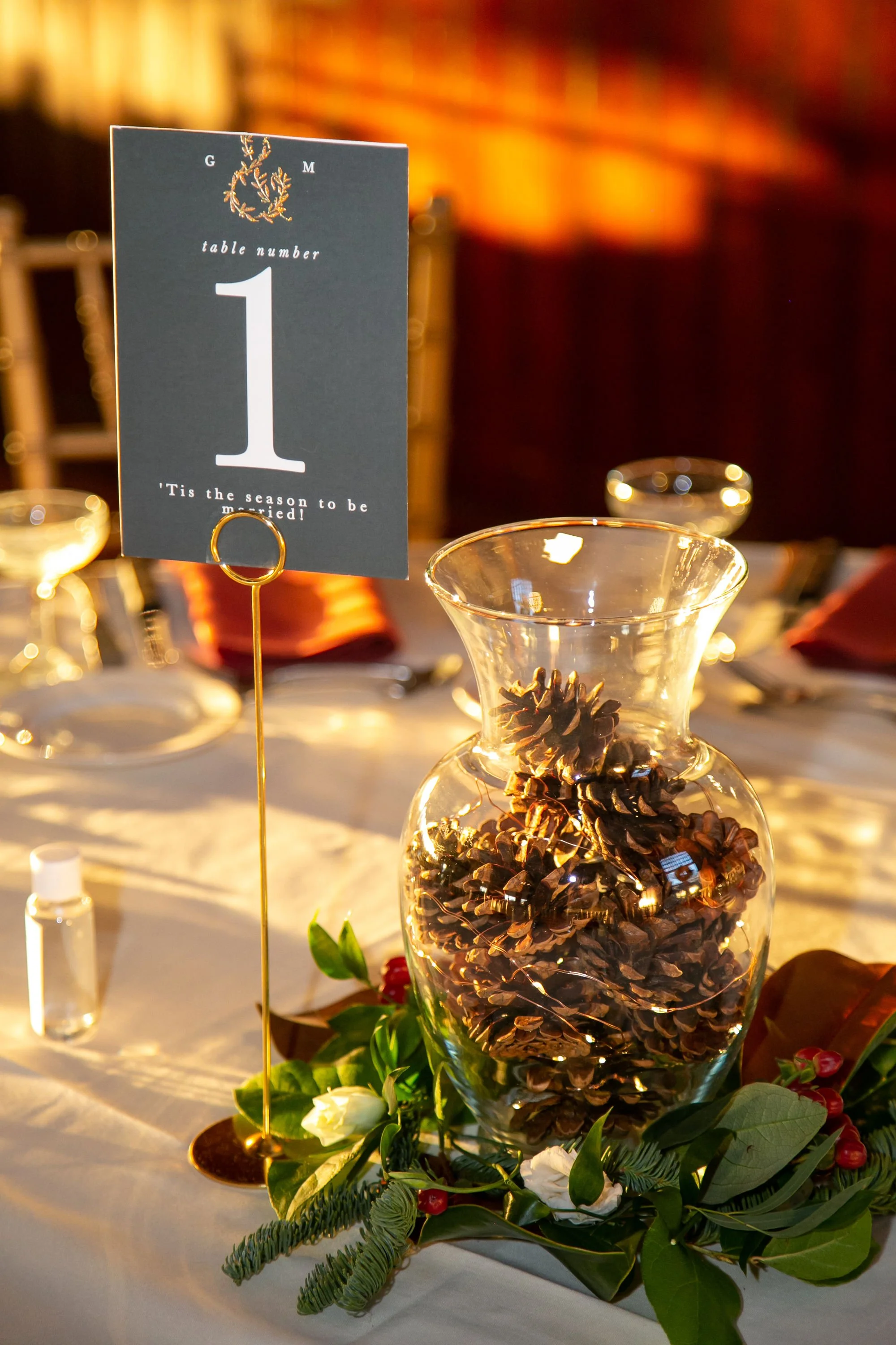 Table centerpiece with a glass jar filled with pinecones, decorated with greenery and flowers, beside a table number sign reading '1' for a wedding reception.