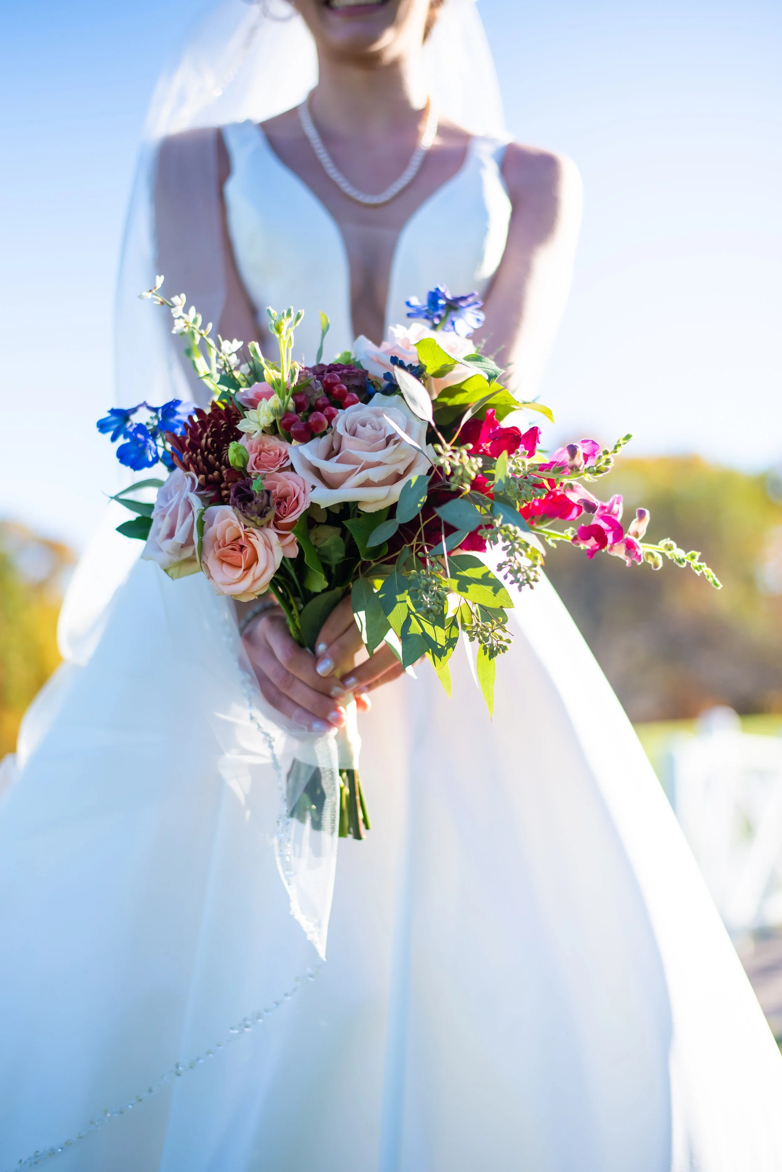 A bride in a white wedding dress holding a bouquet of colorful flowers outdoors on a sunny day.