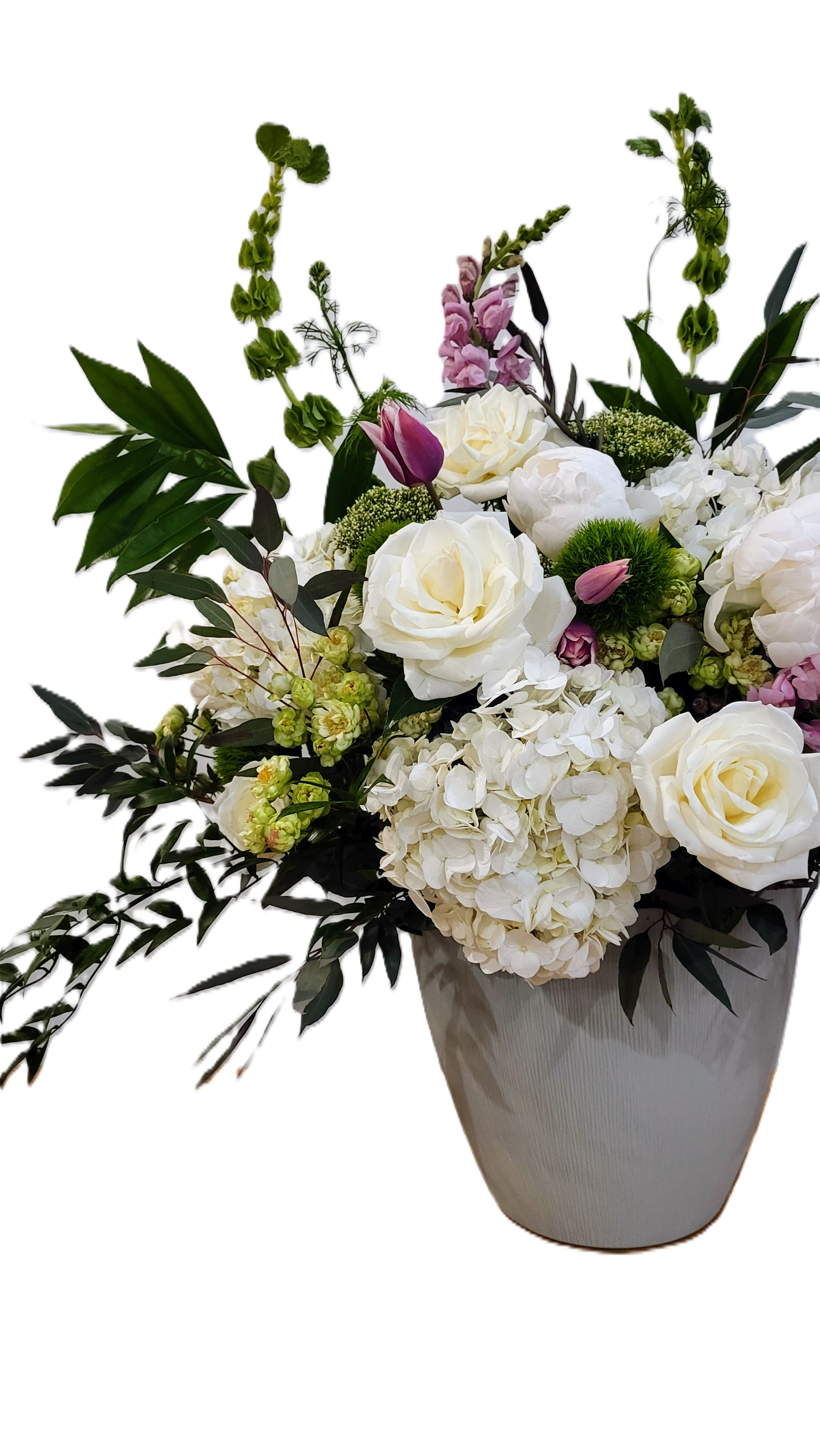 A colorful flower arrangement with pink roses, white hydrangeas, purple delphiniums, pink dahlias, and greenery, placed on the ground beside a wooden fence.