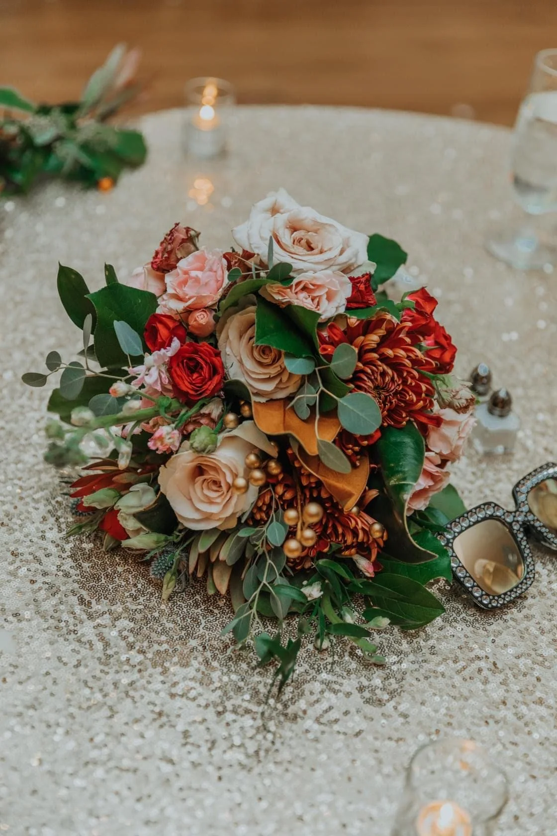 A bouquet of pink, red, and peach flowers with greenery on a glittering tablecloth at a celebration, surrounded by glasses and small decorative objects.
