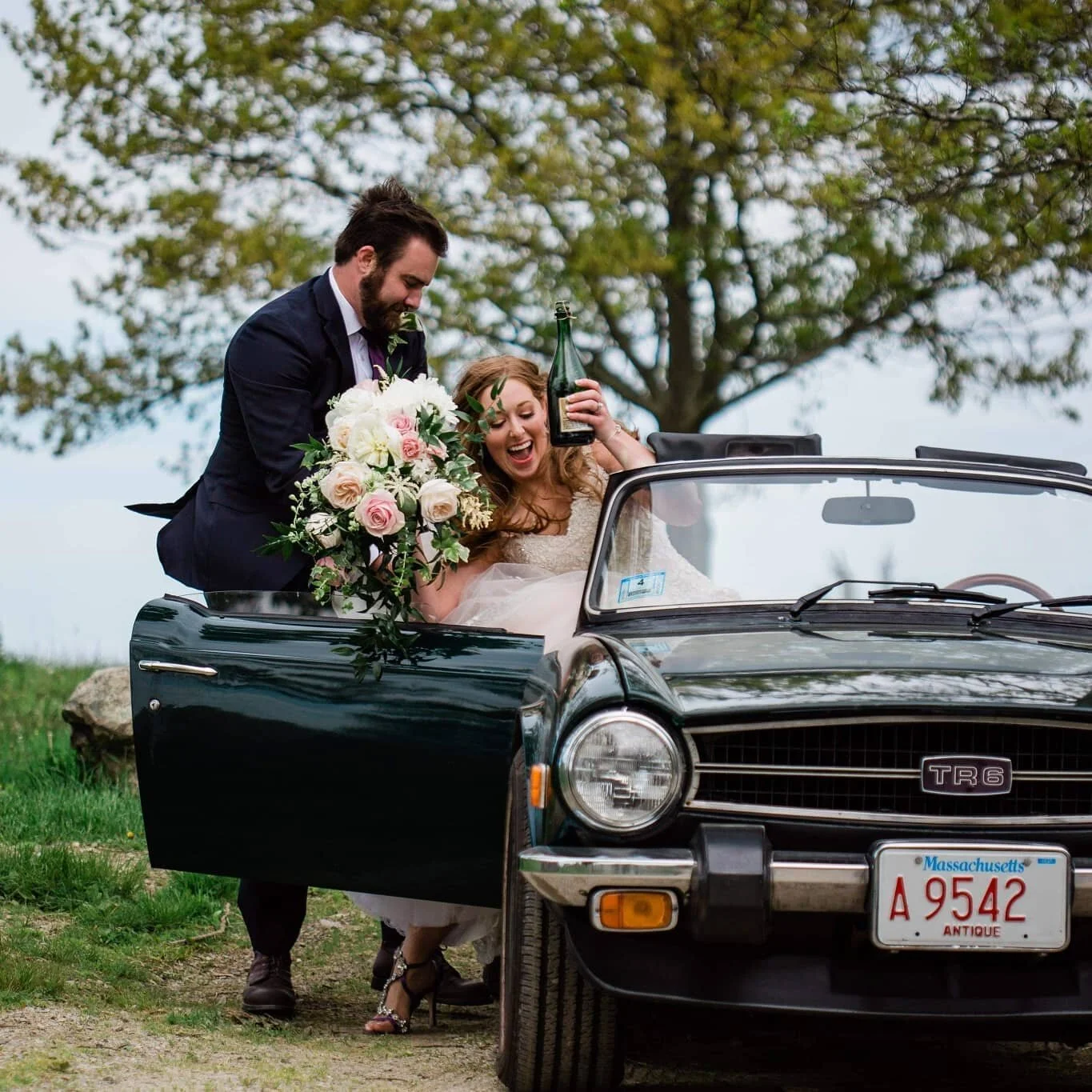 A bride and groom celebrating next to a vintage black sports car, with the bride holding a bouquet of flowers and a champagne bottle, outdoors under a tree.