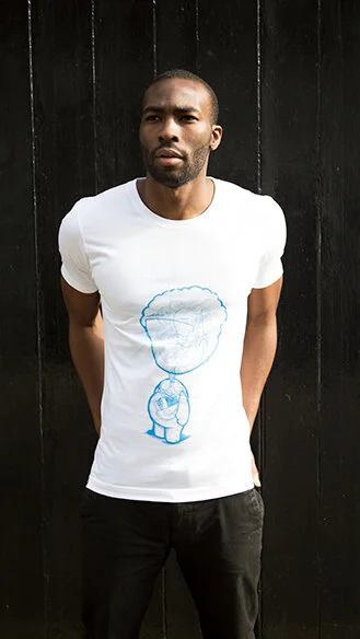 Man standing in front of a black wooden wall, wearing a white t-shirt with a cartoon character design, arms behind his back, looking at the camera.