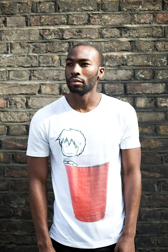 A man with a beard and short hair standing in front of a brick wall, wearing a white T-shirt with a cartoon graphic of a face and a red cup.