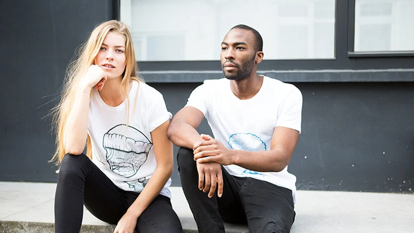 A young woman and man sitting on the sidewalk outside a building, both wearing white T-shirts and black pants, looking contemplative.