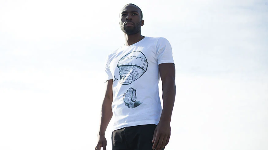 A man standing outdoors against a bright sky, wearing a white t-shirt with graphic designs of lips and a tongue.