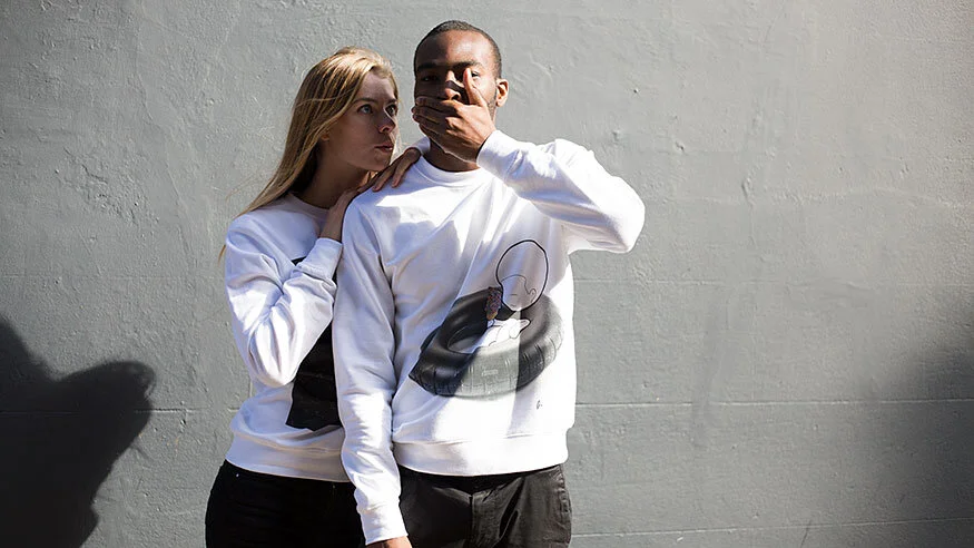 A young woman and man standing against a plain wall, with the woman whispering into the man's ear and the man covering his mouth with his hand, both wearing white sweatshirts.