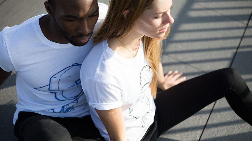 Two people sitting on a concrete surface near a metal railing, with sunlight casting shadows, the woman leaning back with her hands on the ground and the man sitting close behind her.
