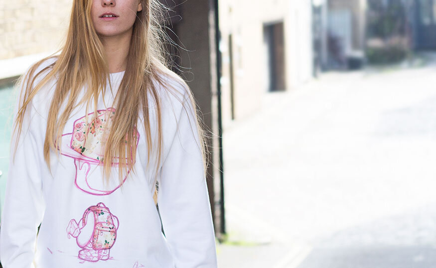 A young woman with long blonde hair wearing a white sweatshirt with colorful cartoon graphics on it, standing outdoors on a city street.