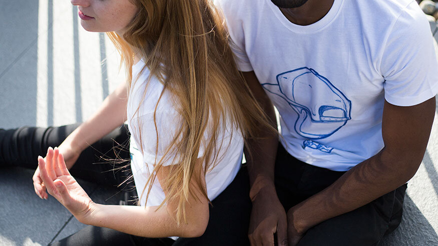 A woman with long red hair practicing yoga or meditation outdoors, sitting against a gray wall, with a man with dark skin and wearing a white graphic t-shirt sitting beside her.
