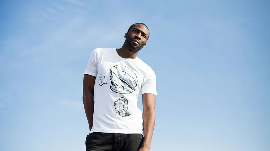 A man standing outdoors against a clear blue sky, wearing a white T-shirt with animated mouth and speech bubble illustrations.