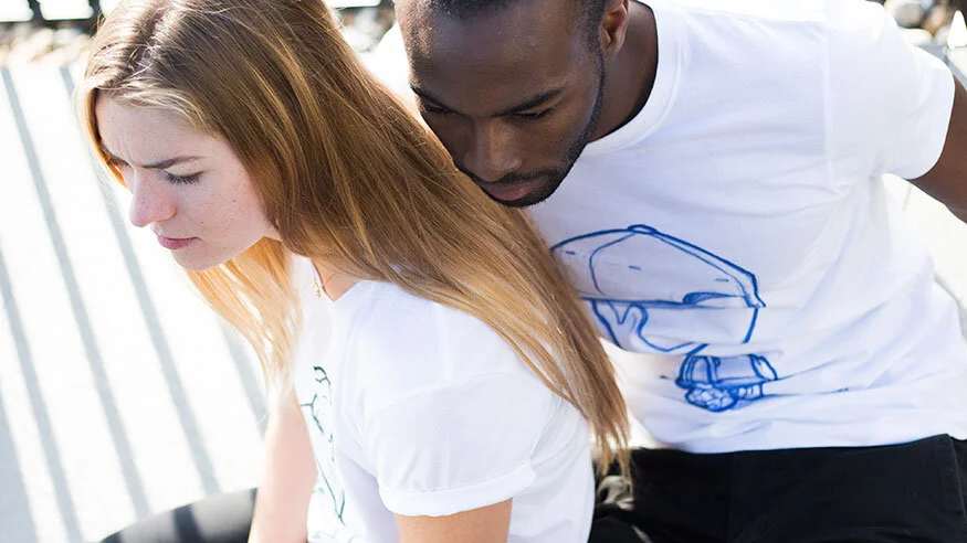Two people, a woman with long red hair and a man with short dark hair, are sitting close together outdoors. They are wearing casual white T-shirts with blue illustrations and appear to be examining or focusing on something out of the frame.