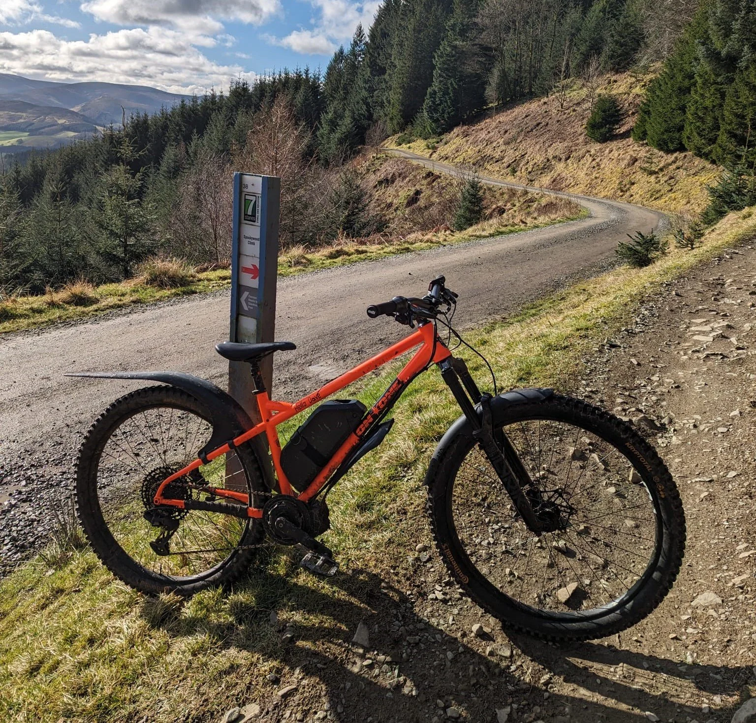 Orange electric mountain bike leaning against a signpost near a gravel mountain trail, with a winding dirt path and forested hills in the background under partly cloudy skies.