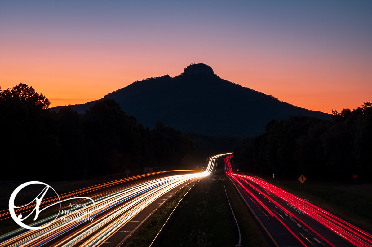 Pilot Mountain photographed after sunset with Highway 52 leading into the distance. Long exposure photograph with light trails created from passing cars.