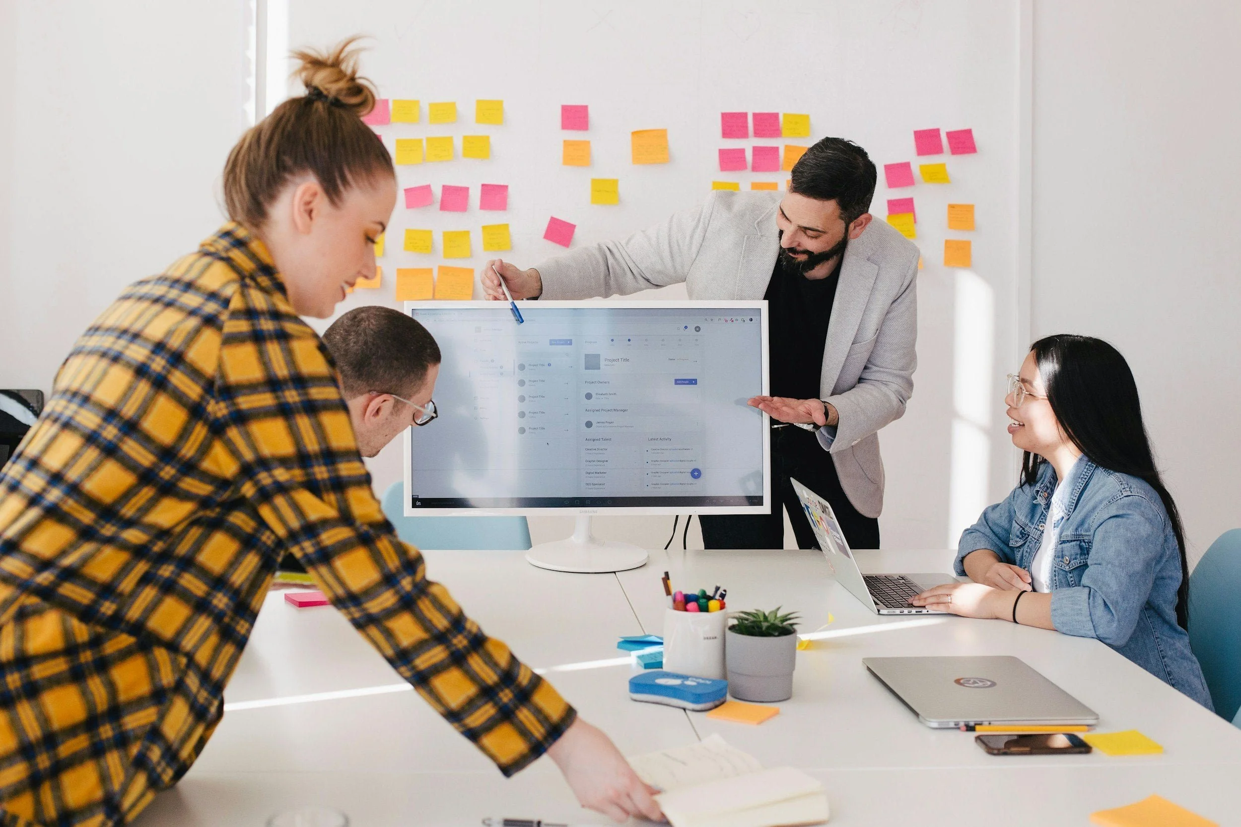 Group of four people in a meeting room with sticky notes on the wall, working around a large desktop monitor, with one person standing and presenting while others are seated and engaged.