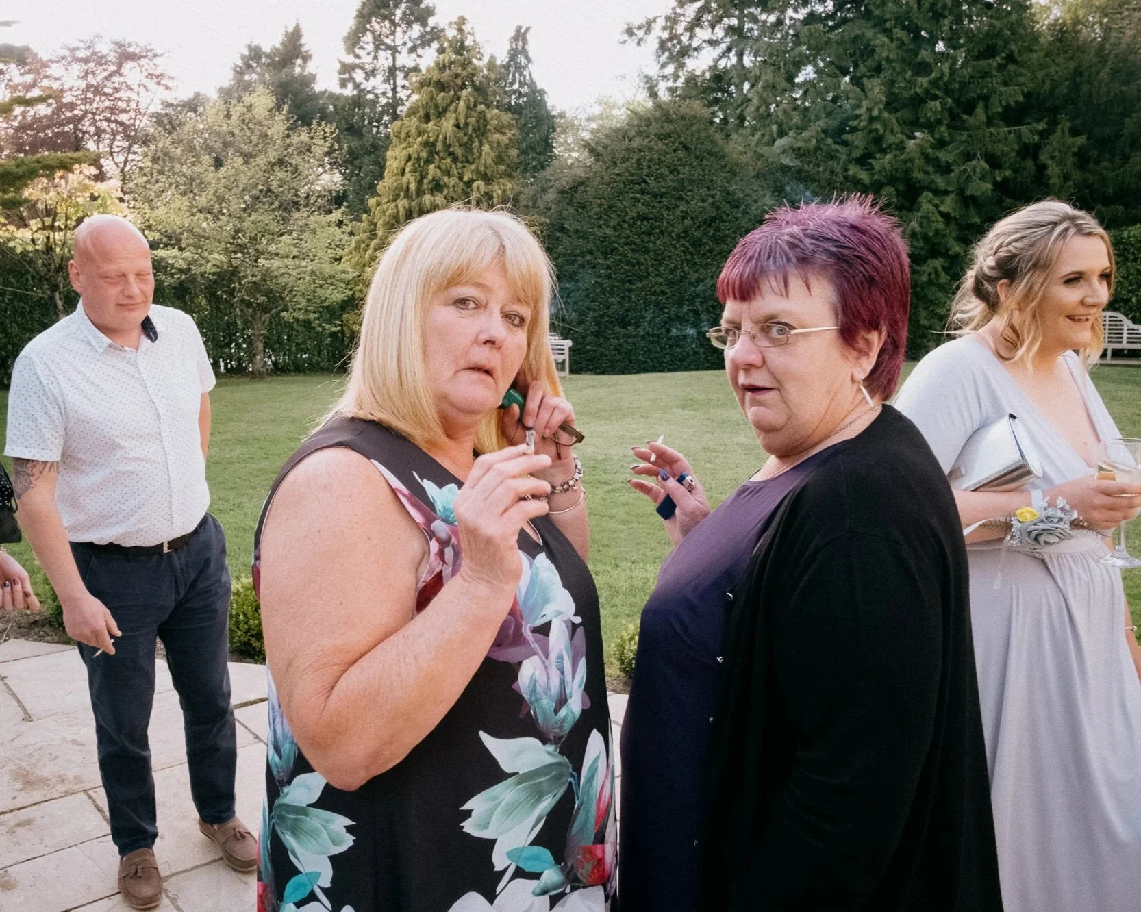Group of four people standing outdoors in a garden, some holding drinks, appearing to be at a social event or gathering.