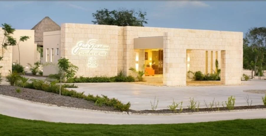 Modern building with illuminated entrance and sign that reads 'Jez Garman Academy of Golf', landscaped with trees, bushes, and a rocky area, and a circular rock with a golf logo in front.