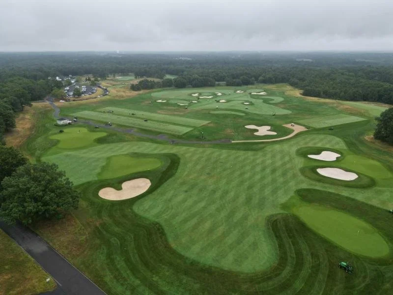 Aerial view of a golf course with fairways, sand traps, and greens surrounded by trees on a cloudy day.