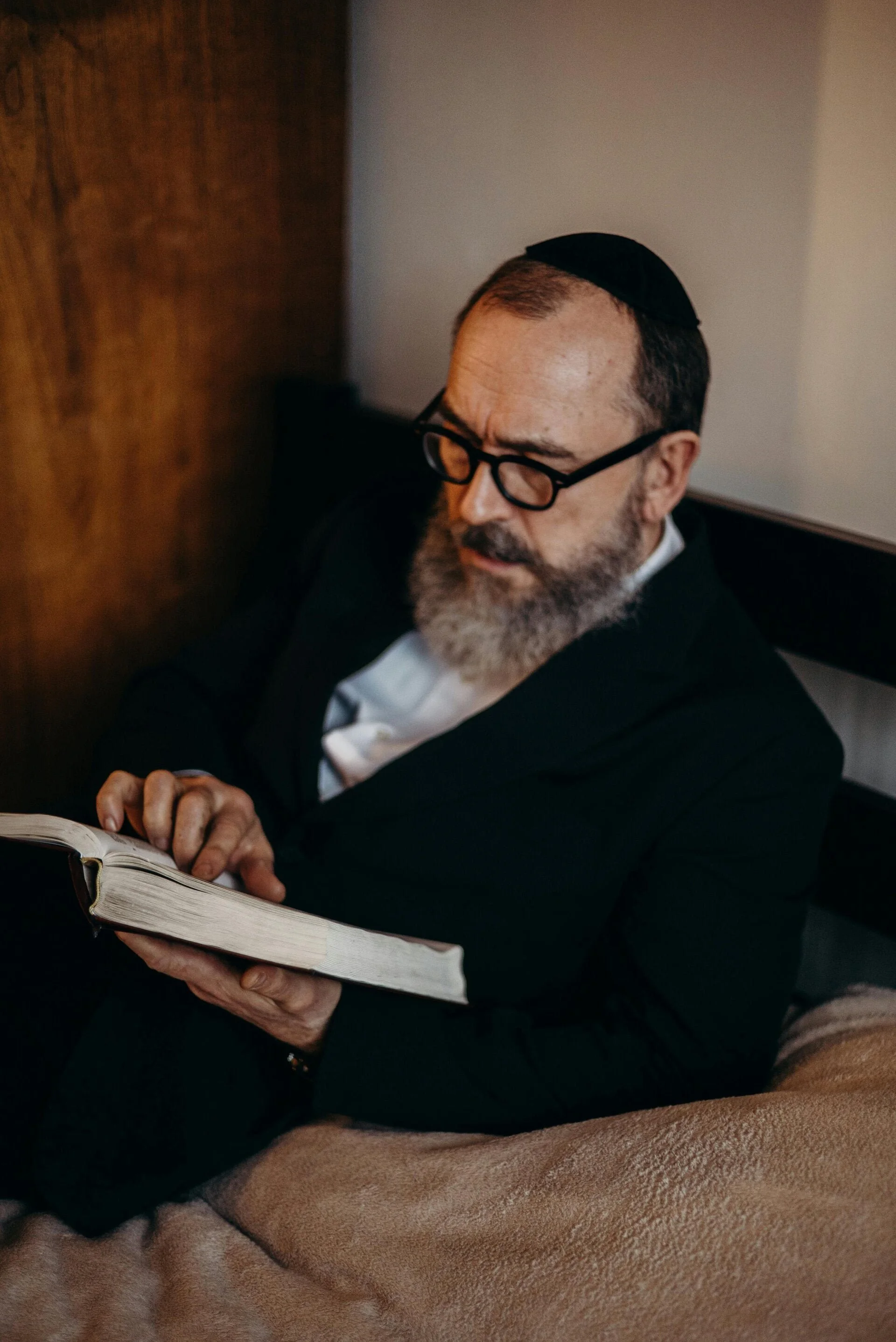 A man with glasses and a beard reading a book while sitting on a bed.