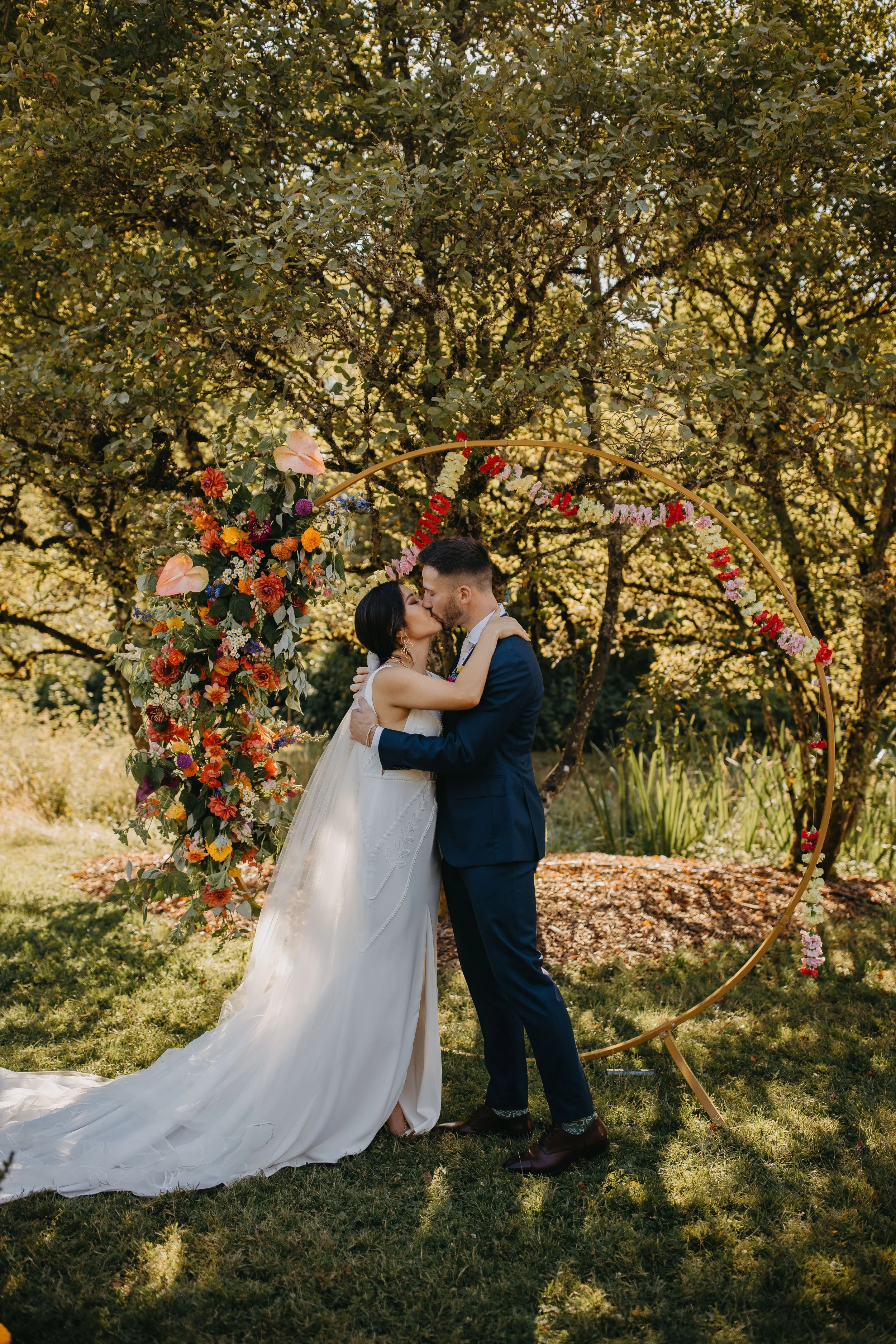 A bright, whimsical ceremony arch with a marigold and carnation garland, bright dahlias, anthurium, and roses on a circular arch in an orchard.