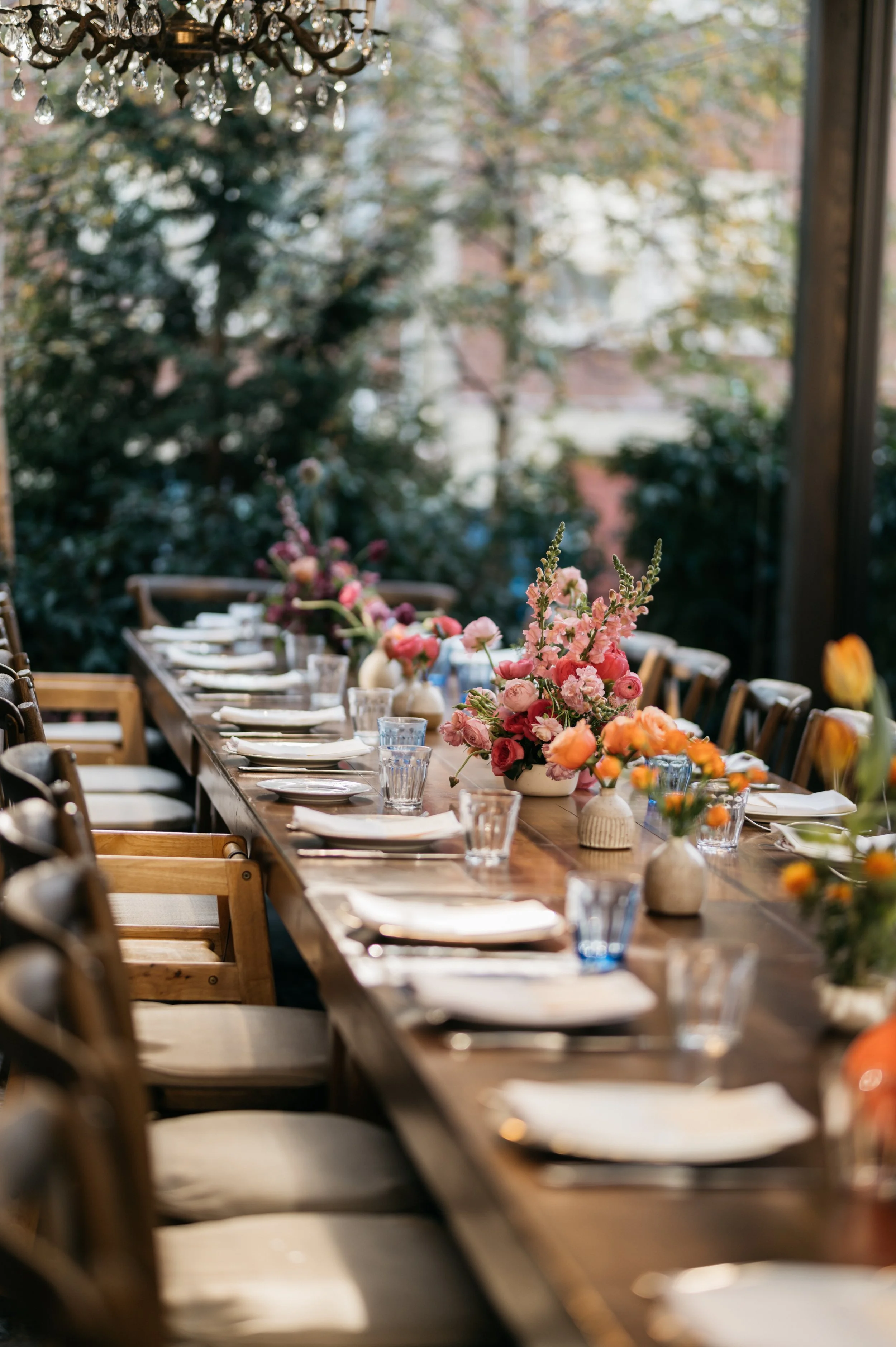 Colorful arrangements in custom ceramic vases, arranged in a row of ombre colors down a wooden table, featuring Spring ranunculus, tulips, and snapdragons.