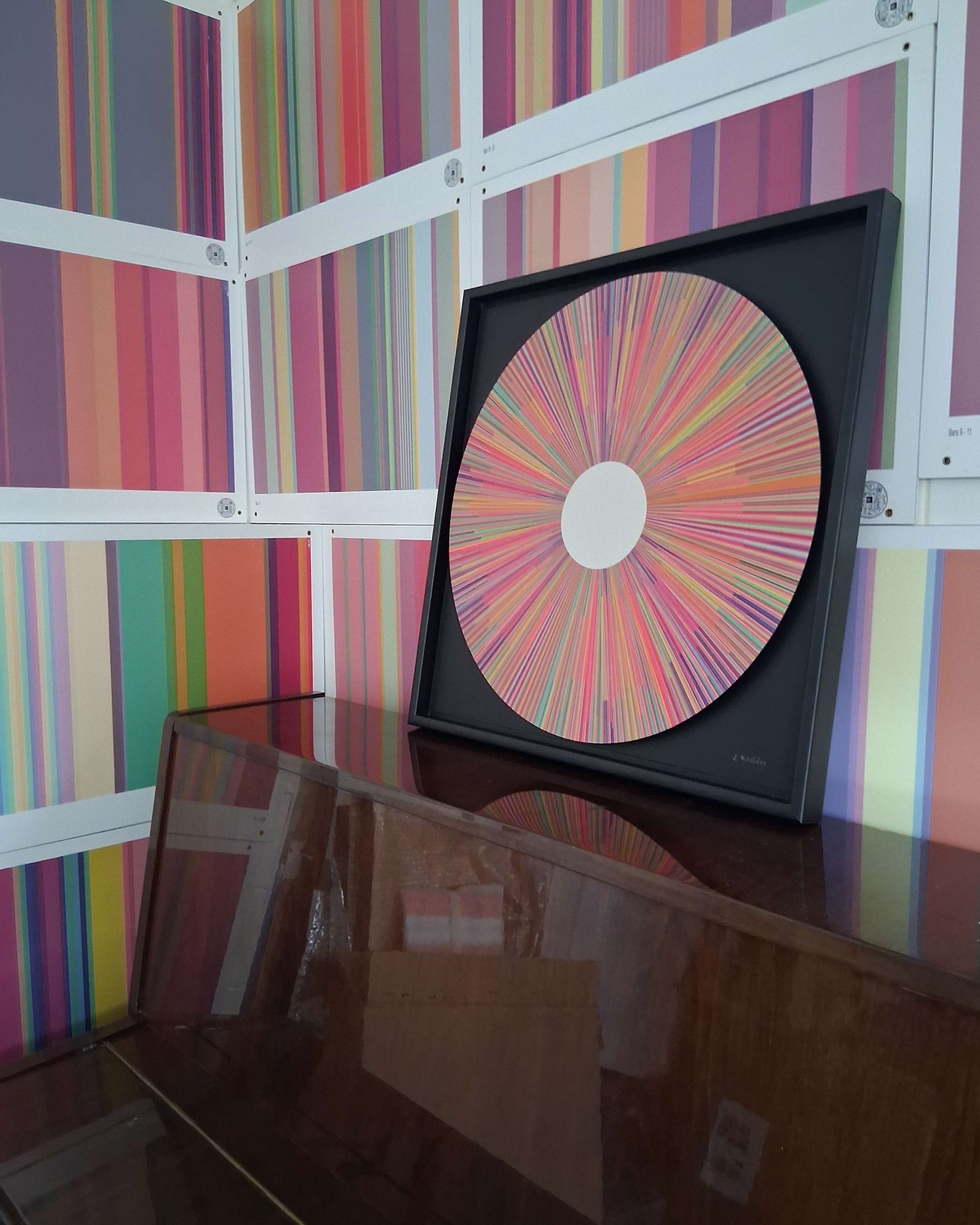 Ave Maria by Elizabeth Mikellides: a circular artwork of radiating colour lines in pinks, oranges and greens, framed in black on an upright piano, with striped colour panels on the studio wall behind.