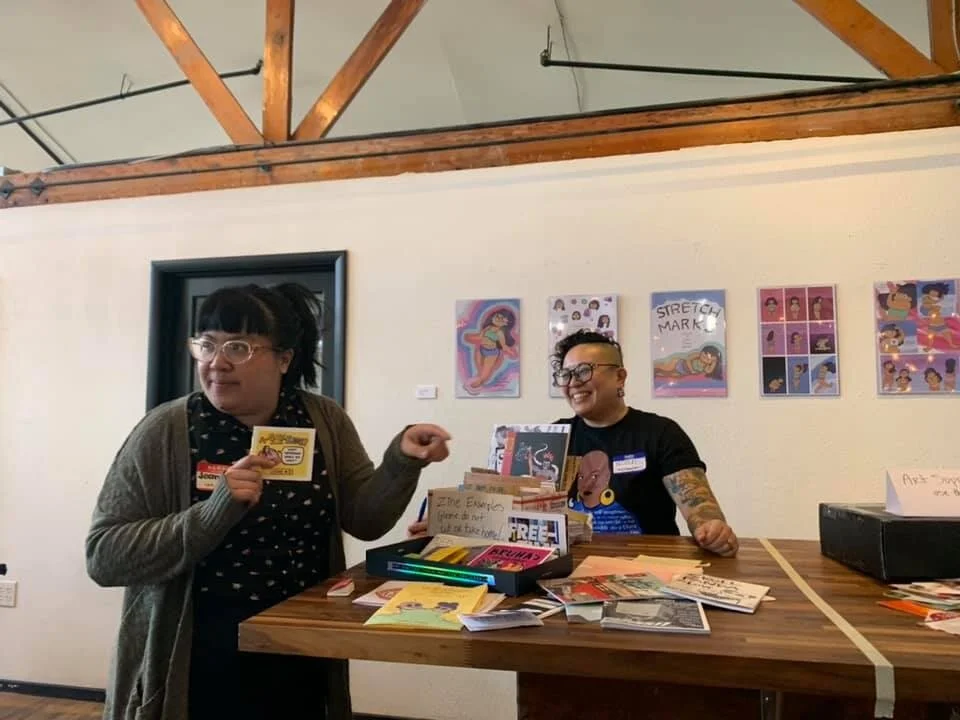  Jean and Nicole stand at a table with many zines. Jean is holding up a zine and talking to someone out of frame.  