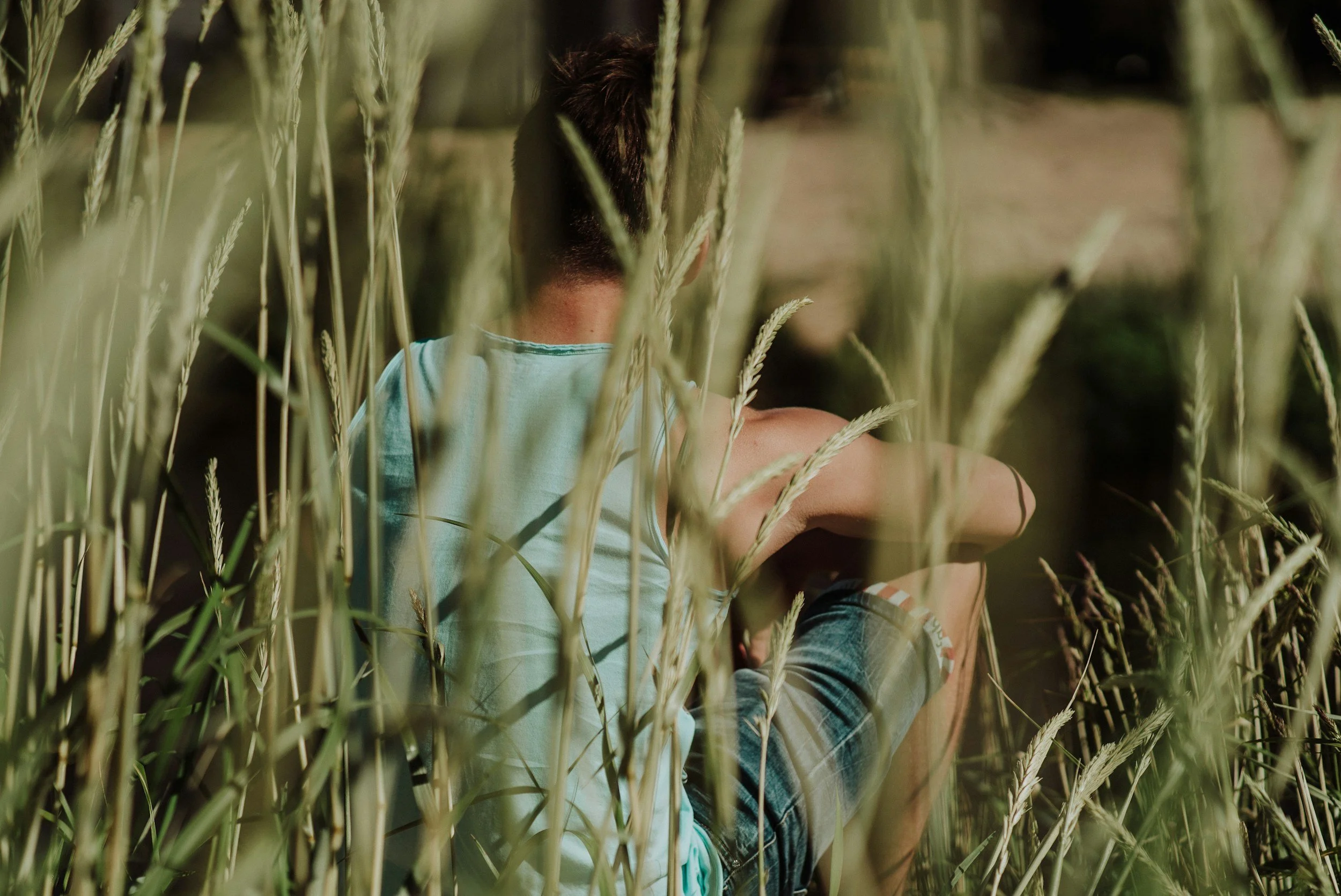 Person sitting in tall grass, wearing a blue tank top and shorts, viewed from behind.