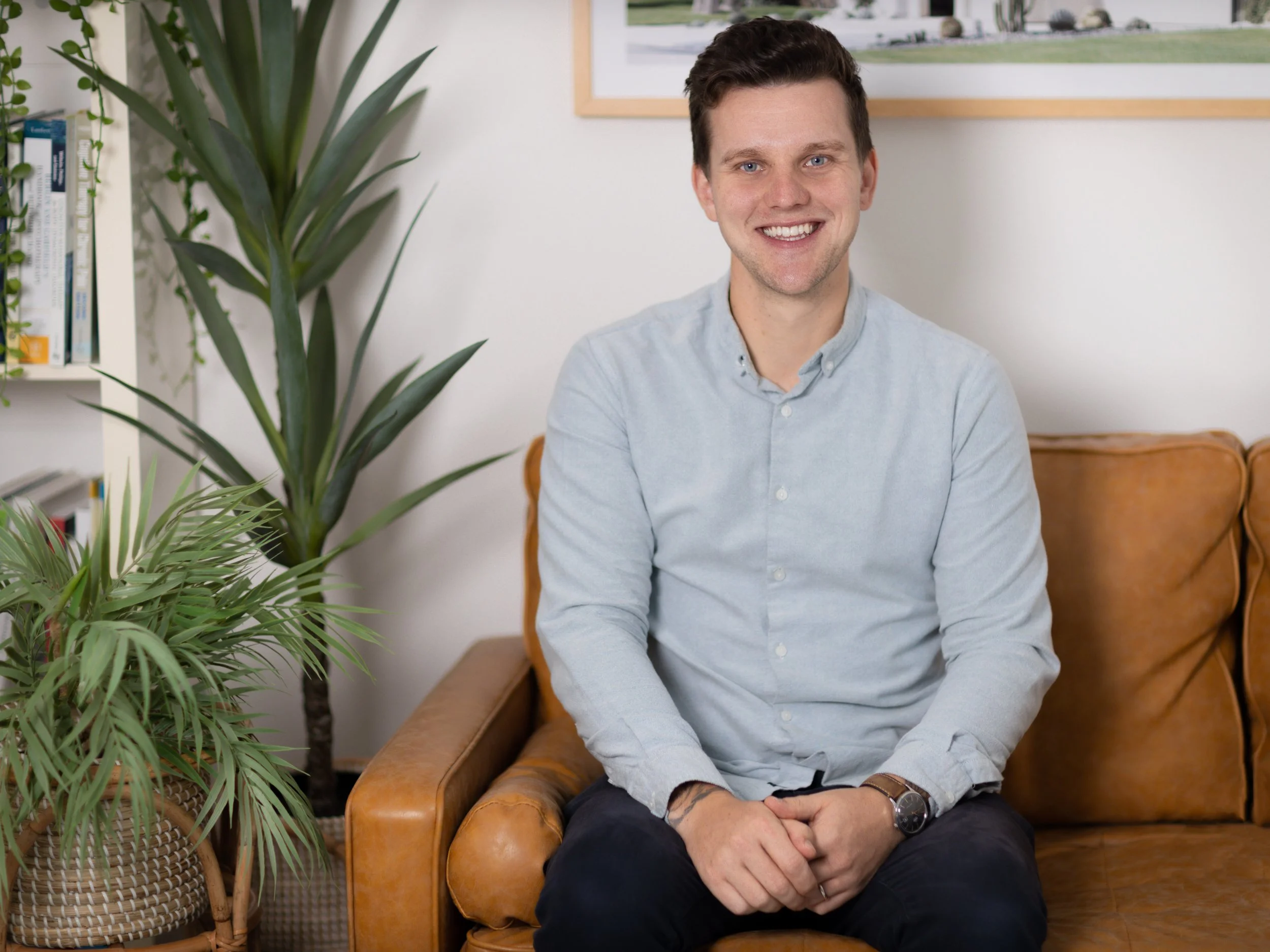 Man sitting on a brown leather couch, wearing a light blue shirt, smiling, with green plants and books in the background.